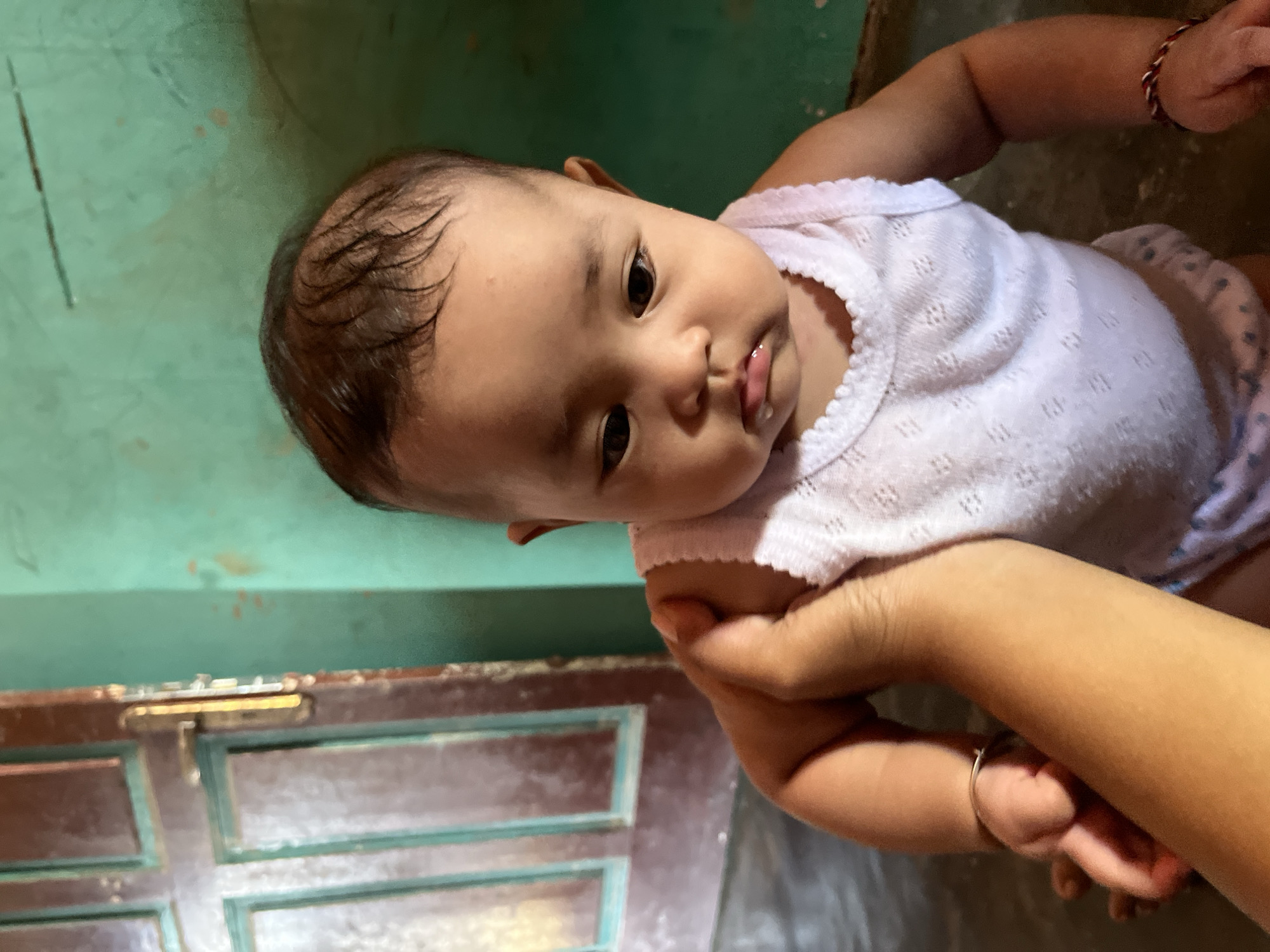 In Kruengmate, Indonesia, a close-up shot captures a baby sitting on the floor. The baby is wearing a white tank top with a textured pattern and appears to be around 6-12 months old, with dark hair and eyes. A human hand, likely an adult's, is holding the baby's left arm, gently supporting them. The background features a portion of a wooden door with a metallic handle, painted a reddish-brown with teal accents. To the right of the door is a teal-colored wall with some subtle markings or dirt. The lighting suggests an indoor environment, possibly a home. The baby's facial expression is neutral, with a slight pout.