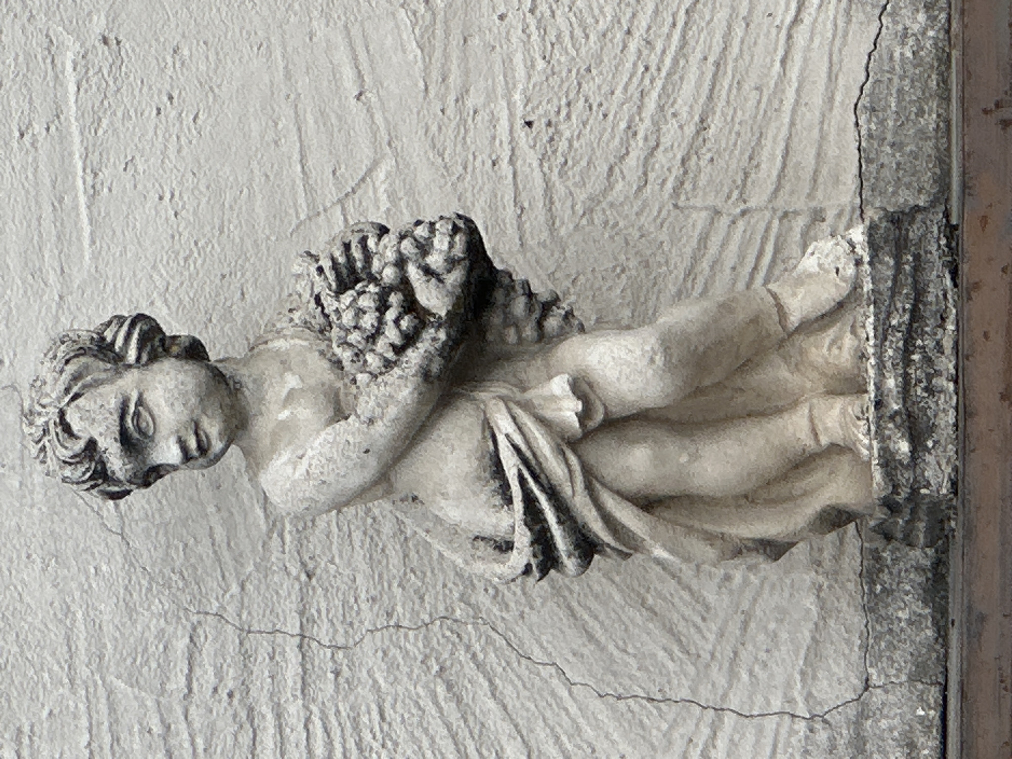 A weathered, light-colored stone statue of a child, possibly a cherub, stands against a rough, white textured wall. The statue depicts the child in a classical pose, draped in cloth, and holding a bunch of grapes in its arms. The stone is worn and shows signs of age and exposure to the elements, with some discoloration and minor cracks. The child's face is rendered with soft features, looking slightly to the left. The background wall exhibits a coarse, stucco-like finish with visible horizontal lines and a few vertical cracks. The statue is positioned on a grey, granular base, which appears to be made of concrete or a similar material, and is slightly chipped and worn. The bottom of the image shows a dark horizontal surface, possibly a railing or ledge.