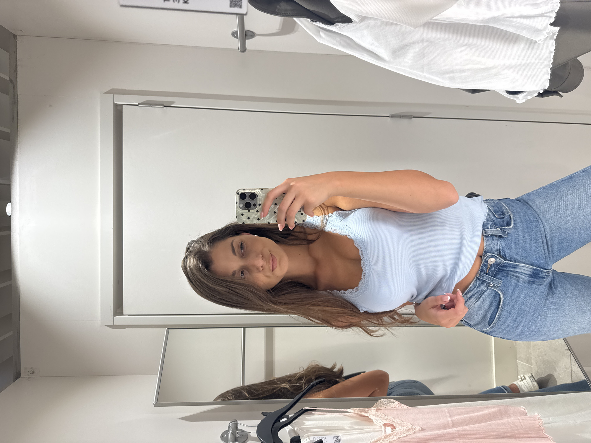 A young woman is taking a selfie in what appears to be a dressing room. She is wearing a light blue tank top with lace trim and blue jeans. She is holding a smartphone with a dotted case. The dressing room has a mirror, white walls, and a clothing rack with white and pink garments. The location is likely a clothing store, but there is not enough detail to determine if it is in Burgas, Bulgaria.
