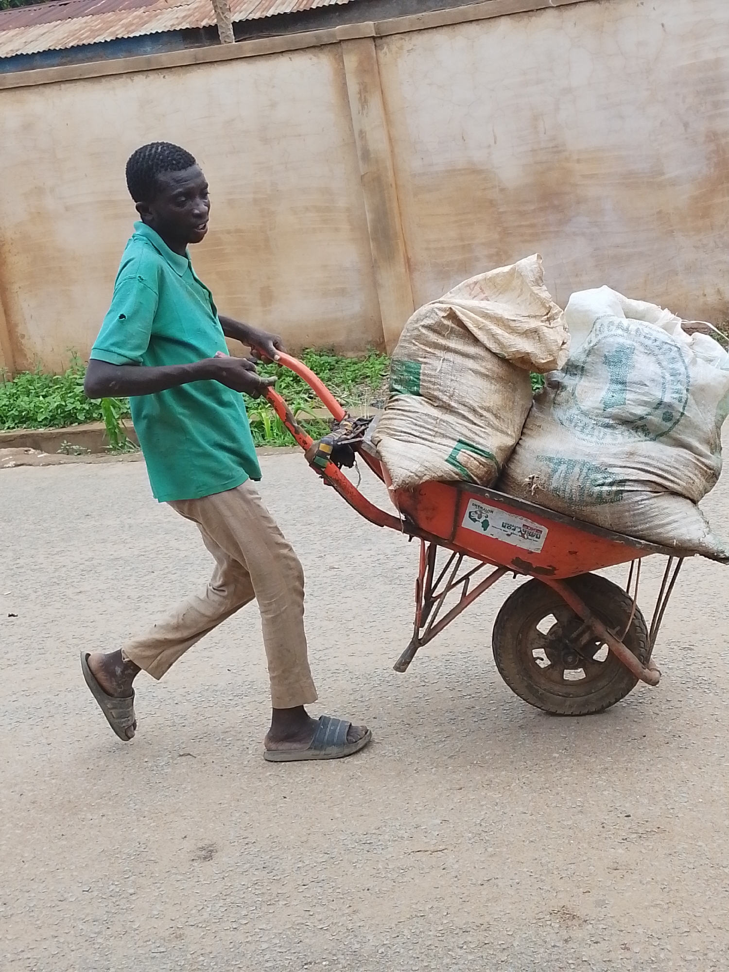 A young man is seen pulling an orange wheelbarrow loaded with two large, full sacks on an asphalt road. He is wearing a light green polo shirt, tan trousers, and slides. The location appears to be in Jos, Nigeria. A low concrete wall is in the background, with a building and some greenery visible behind it.