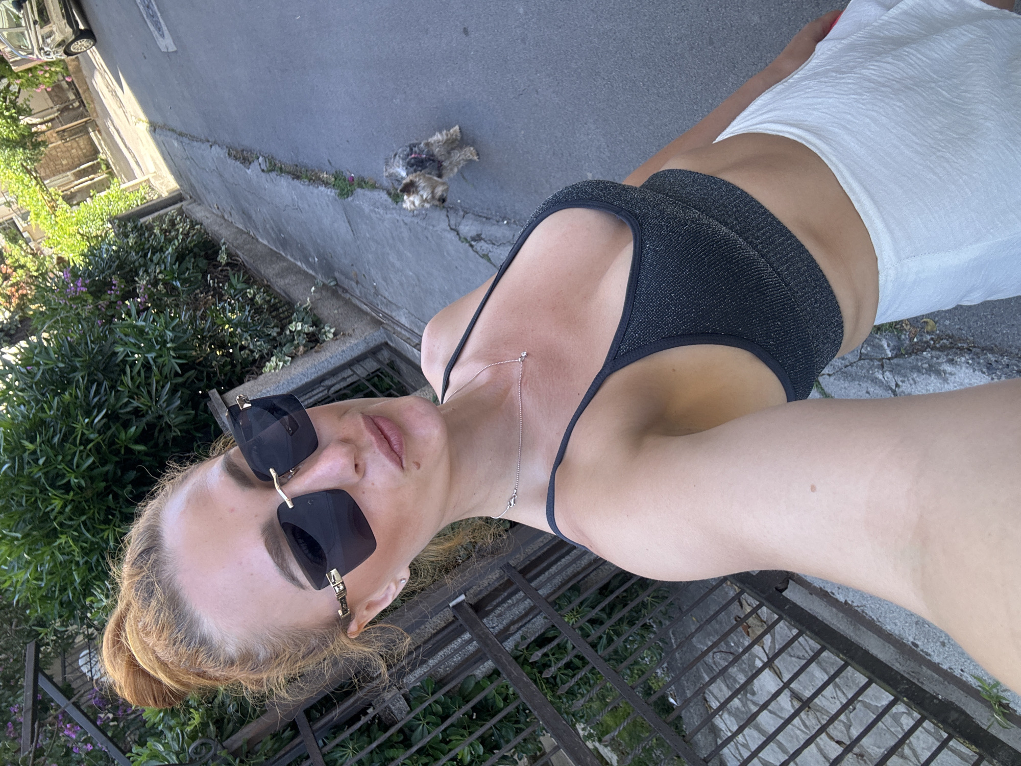 In Budva, Montenegro, a woman with red hair pulled into a bun, wearing sunglasses, a sparkly black crop top, and a white skirt, takes a selfie. Her head is angled slightly down and to the side. A small dog stands on the paved street behind her. Green foliage and a metal fence are visible to her left, and a parked car is further down the street to the right. The scene appears to be a residential street with a sidewalk.