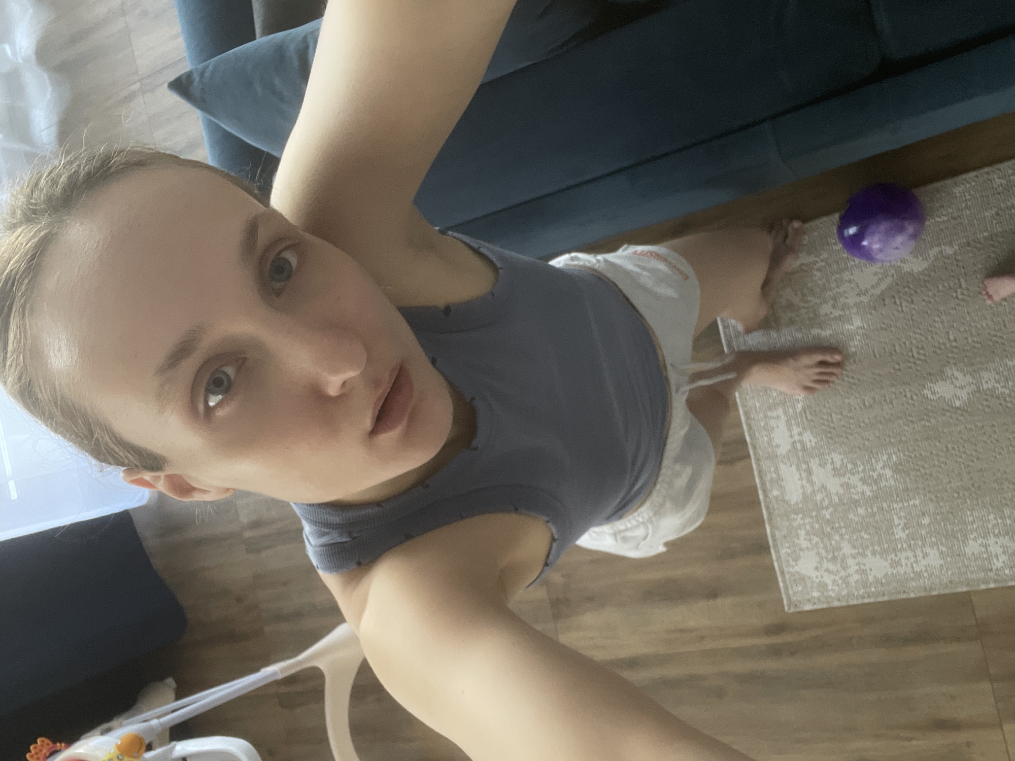 A woman takes a selfie indoors. She wears a grey tank top and white shorts. A blue couch is partially visible in the background. A purple ball sits on a beige rug on a wood floor. The location is not discernible as being in Подшивалово, Russia based on the image. A white baby walker is also visible.