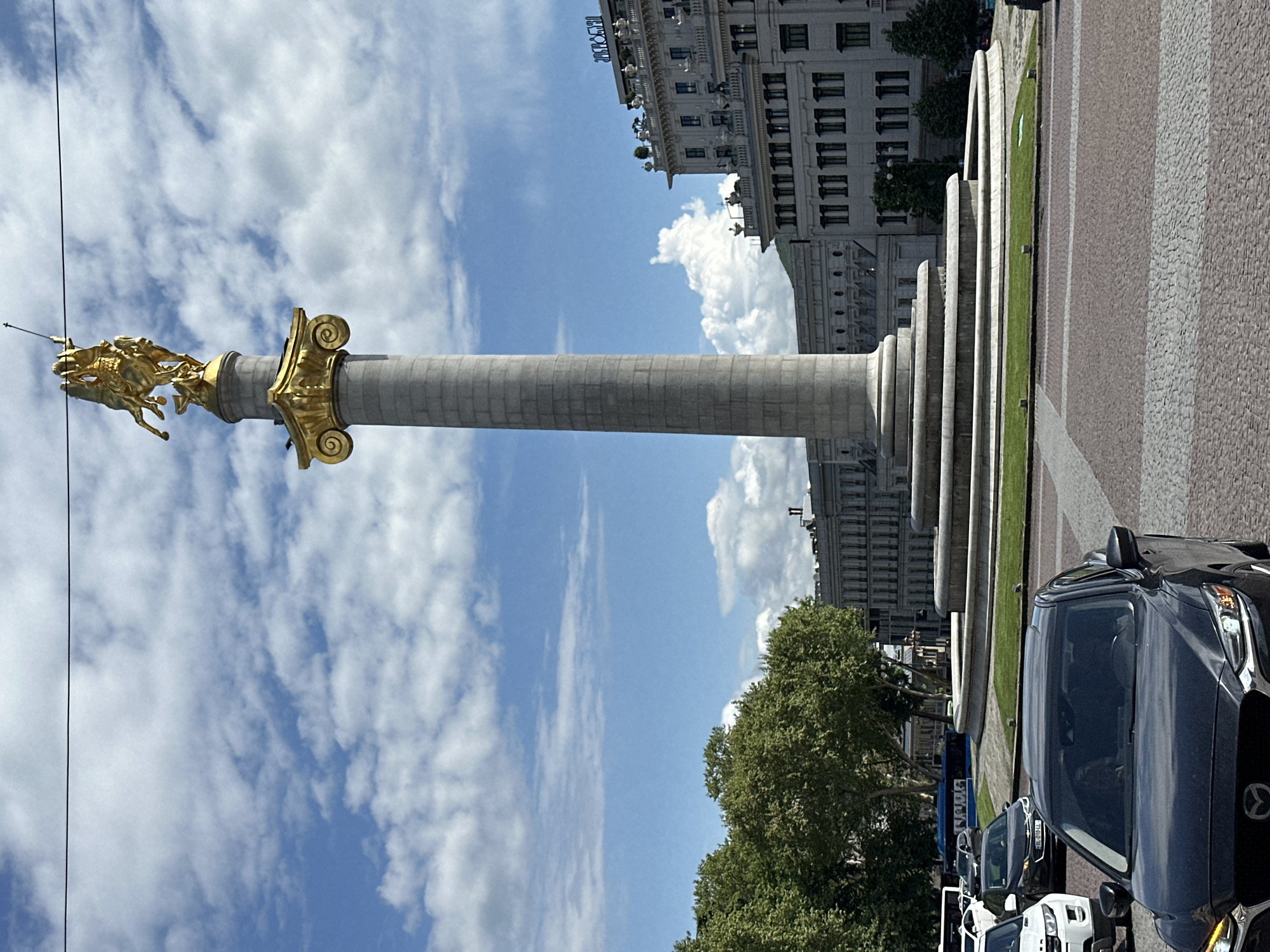 The image shows the St. George Monument in Tbilisi, Georgia, against a cloudy sky. The monument consists of a tall stone column topped with a golden statue of St. George slaying a dragon. The column is set on a tiered base. Nearby is a multi-story building and a row of parked cars.