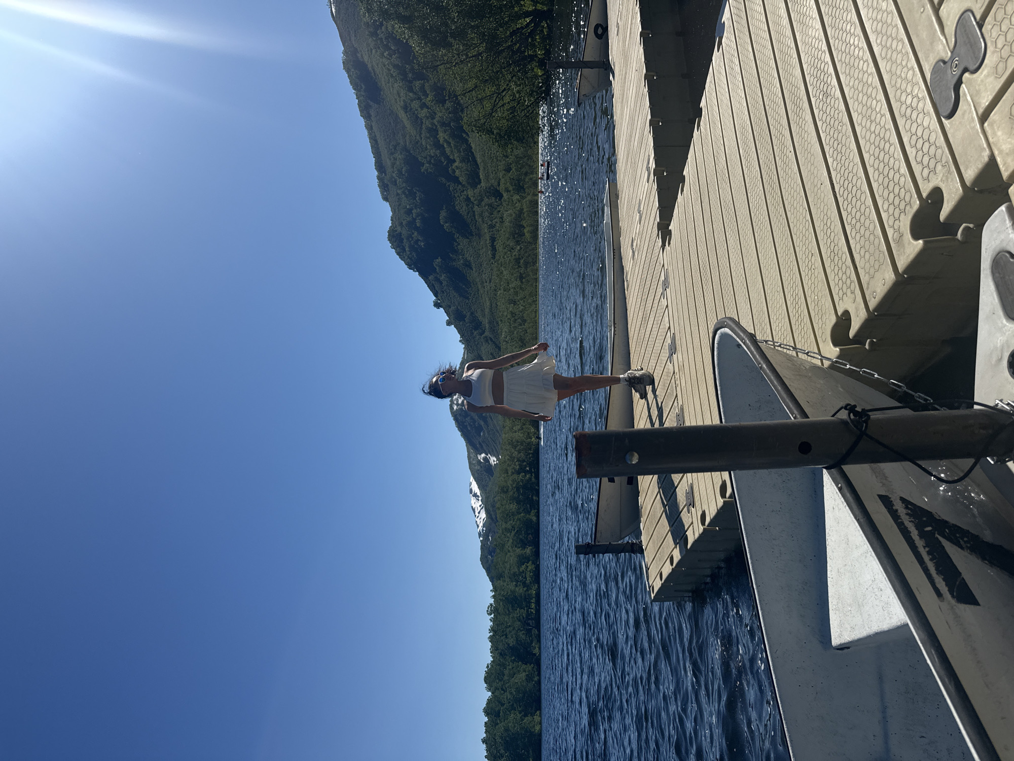 The image captures a sunny day at a lake, likely in or near Paratunka, Russia, given the presence of mountains and lush green vegetation. In the foreground, a person, appearing to be a woman, is walking on a wooden dock. She is wearing a white crop top and a white skirt, with sunglasses. The dock is constructed of modular, light-colored plastic sections and extends out over the dark blue water. Two small boats are moored alongside the dock. The lake is calm, with gentle ripples on the surface, and the sun glints off the water. In the background, a tree-covered mountain range rises, with patches of snow visible on its higher peaks. The sky is a clear, bright blue with a lens flare from the sun in the upper right corner. The scene suggests a leisurely outdoor activity, possibly in a resort area, given the amenities like the dock and boats.