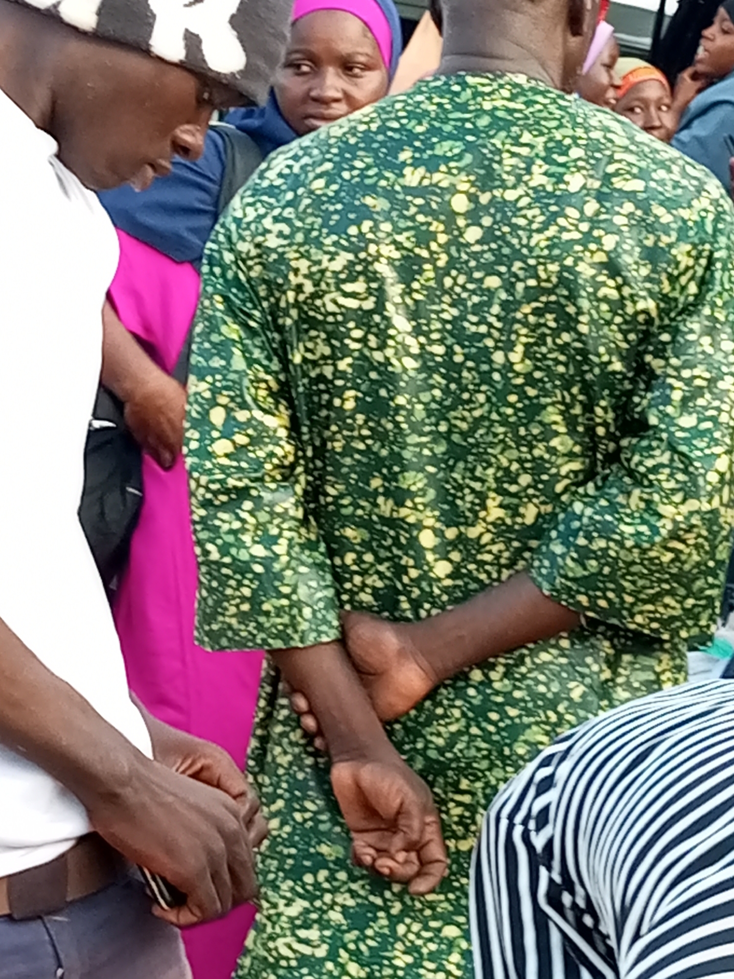 The image depicts a group of people in an outdoor urban setting in Gombe, Nigeria. In the foreground, a person wearing a white t-shirt and a patterned beanie is visible from the side, with their head bowed and hands clasped in front. To their right, another person wears a vibrant pink garment. Central to the image is an individual in a green, yellow-speckled patterned top with their back to the camera, their arms clasped behind them. Several other individuals are partially visible behind them, some wearing headscarves in shades of pink and orange, and one person in a blue hooded garment. The background is blurred, suggesting a busy environment, and the lighting indicates it is daytime.