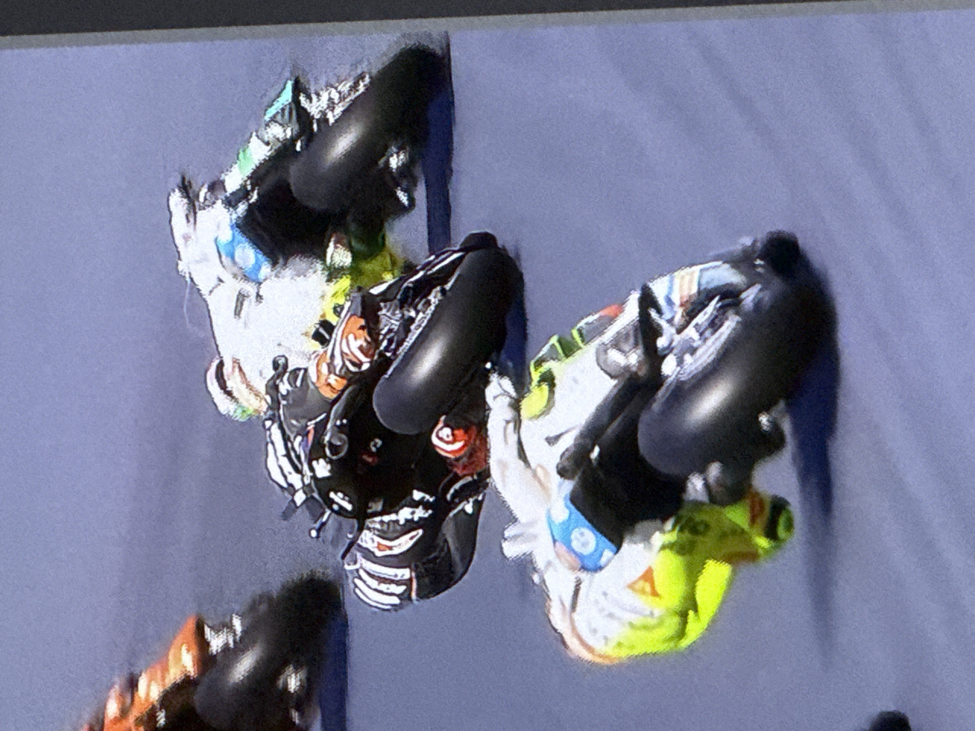 The image shows a motorcycle race with three motorcycles and their riders in motion on a grey track. The motorcycle in the foreground is predominantly yellow and white with black accents, and the rider is wearing a white helmet with some green and colored detailing. Behind and to the right of this motorcycle is another motorcycle, mostly black with white and red accents, and the rider is wearing a black helmet with white detailing. Further back and to the left, a third motorcycle, colored orange and black, is visible. The image is a photograph taken from a slightly elevated angle, capturing the dynamic movement of the racers. The background is a muted, textured grey, consistent with a racetrack surface. There is no discernible urban environment, buildings, or any other features that would indicate a specific city location. Therefore, it is not possible to determine if this image is located in Cahul, Moldova.