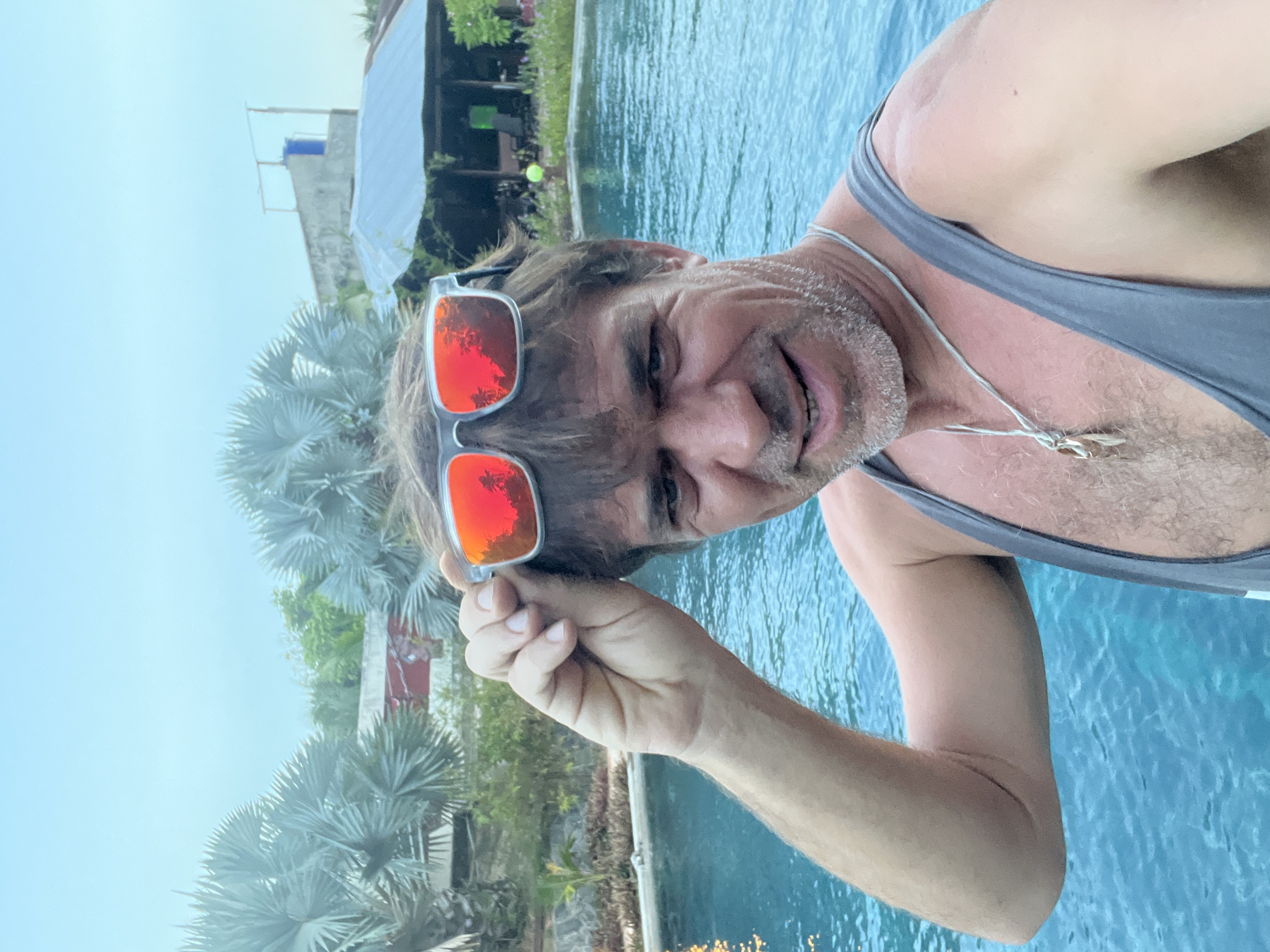 A man wearing a grey tank top and sunglasses is taking a selfie by a swimming pool in Phu Thuy Ward, Vietnam. The sunglasses have red lenses reflecting palm trees. The man has light brown hair and stubble on his face. He is wearing a silver necklace with a pendant. Behind him, the swimming pool has blue water with reflections. Palm trees are visible in the background, along with a building with a tin roof and a blue water tank on top. The sky is overcast and light blue.
