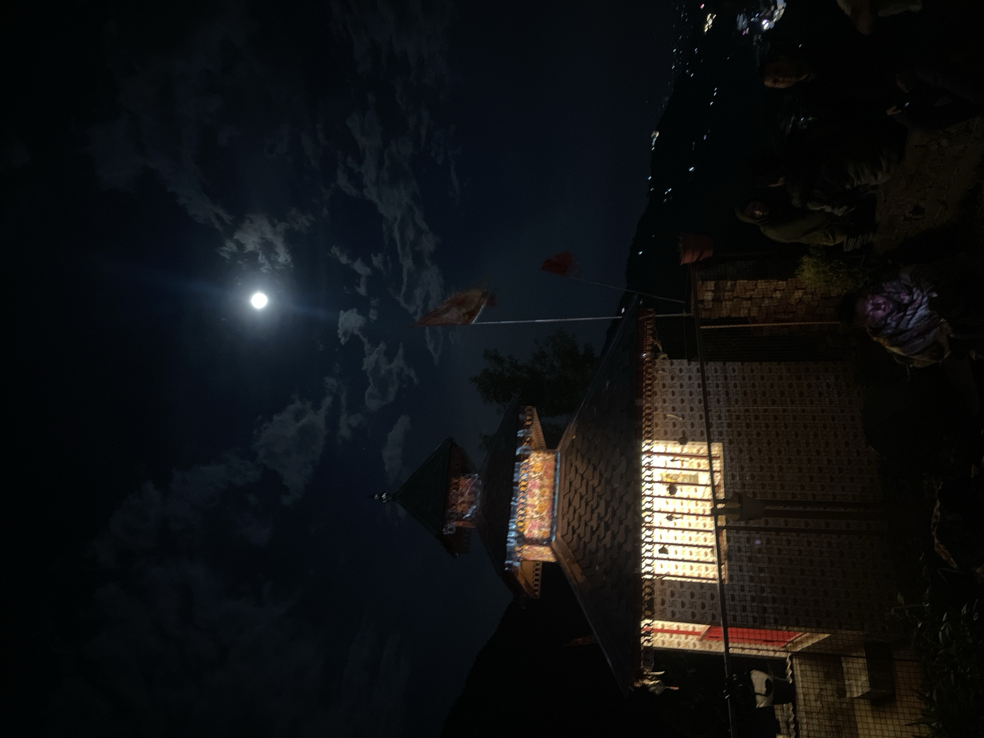 The image depicts a night scene in Haini, India, featuring a building illuminated by a bright light emanating from within. The sky is dark with visible clouds and a full moon. Two flags are attached to poles near the building. In the darker, upper-right corner of the image, several people are visible, some appearing to be wearing hooded clothing. A figure with a patterned head covering is also present near the building. The environment suggests an outdoor setting, possibly near vegetation.