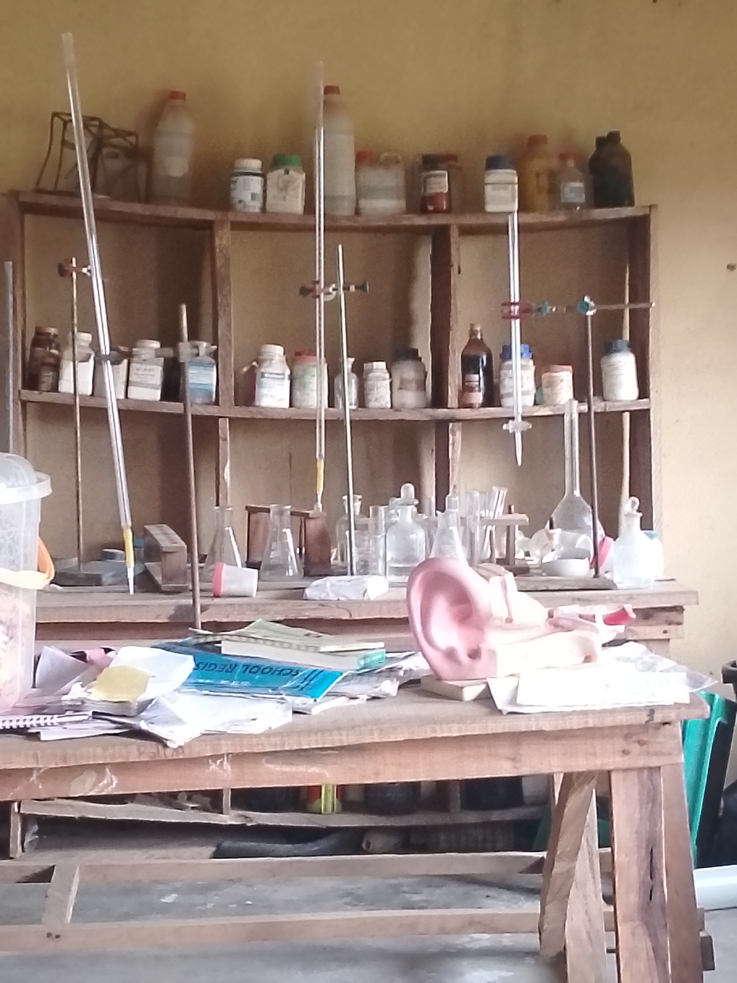 The image shows a disorganized laboratory setup. A wooden shelf unit, likely in a school or community setting, is filled with various scientific equipment and supplies. On the top shelf, several bottles and containers are visible, including what appear to be chemical reagents. The middle shelf holds more bottles and apparatus, such as burettes and stands, suggesting practical experiments are conducted here. The bottom shelf, less visible, also contains items. In front of the shelves, a wooden table is cluttered with papers, some stacked neatly, others scattered. A blue notebook or register labeled "SCHOOL REGIS" is prominent amongst the papers. A pink anatomical model, possibly of an ear, rests on the table, implying educational or medical purposes. The overall environment appears to be indoors, with a plain, light-colored wall in the background. The scene suggests a functional but somewhat neglected laboratory space, possibly in Serti, Nigeria, given the context provided. There are no people visible in the image.