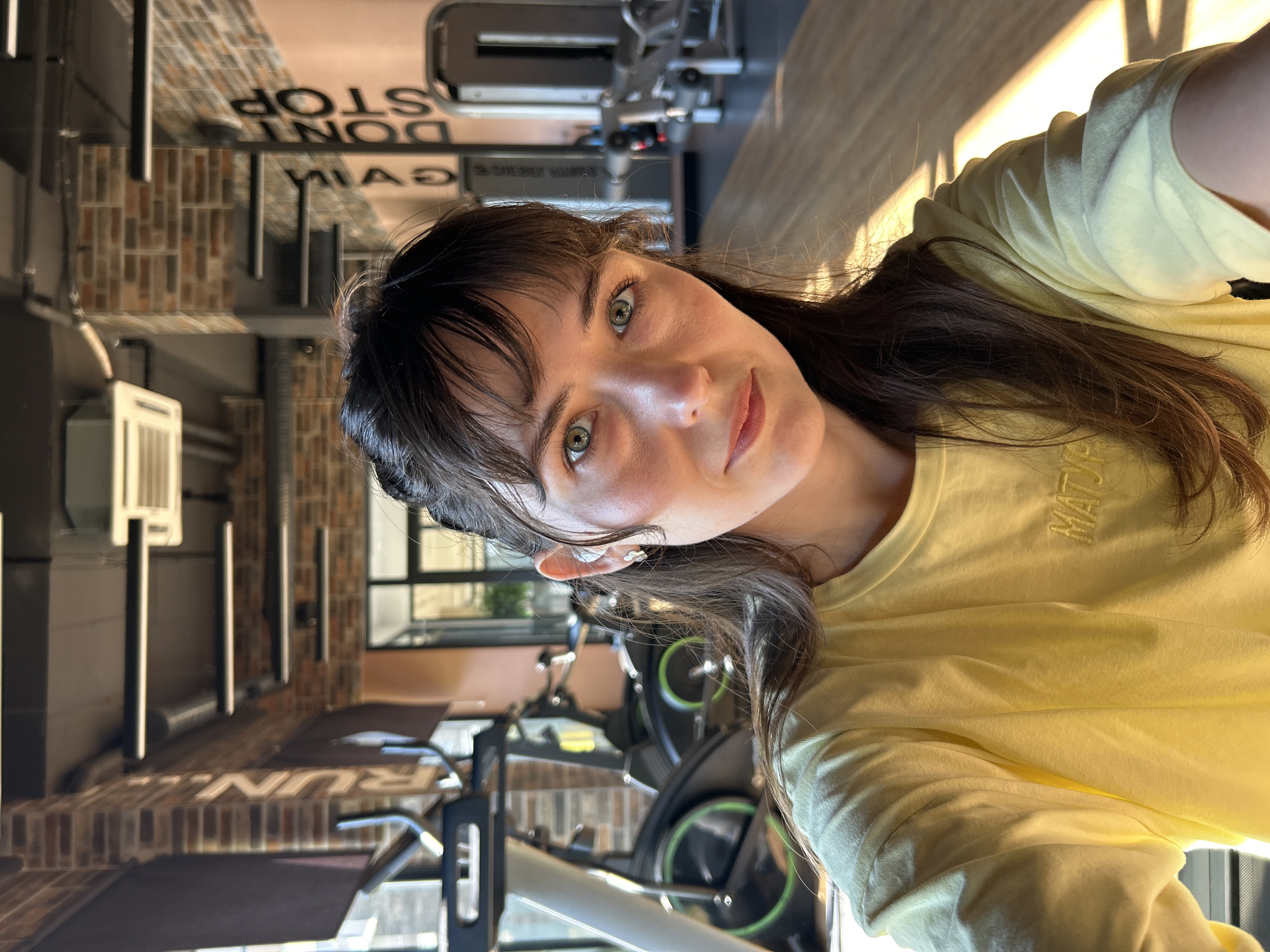 A woman with brown hair and green eyes is posing for a selfie in a gym. She's wearing a yellow t-shirt. The background reveals exercise equipment, including stationary bikes and treadmills, and a brick wall with "DON'T GAIN STOP RUN" printed on it. The gym's interior appears modern with a mix of dark and light colors. While the image doesn't explicitly state the location, based on the presence of Turkish words like "Alanya", and other contextual clues, it is highly probable that the gym is located in Alanya, Turkey.