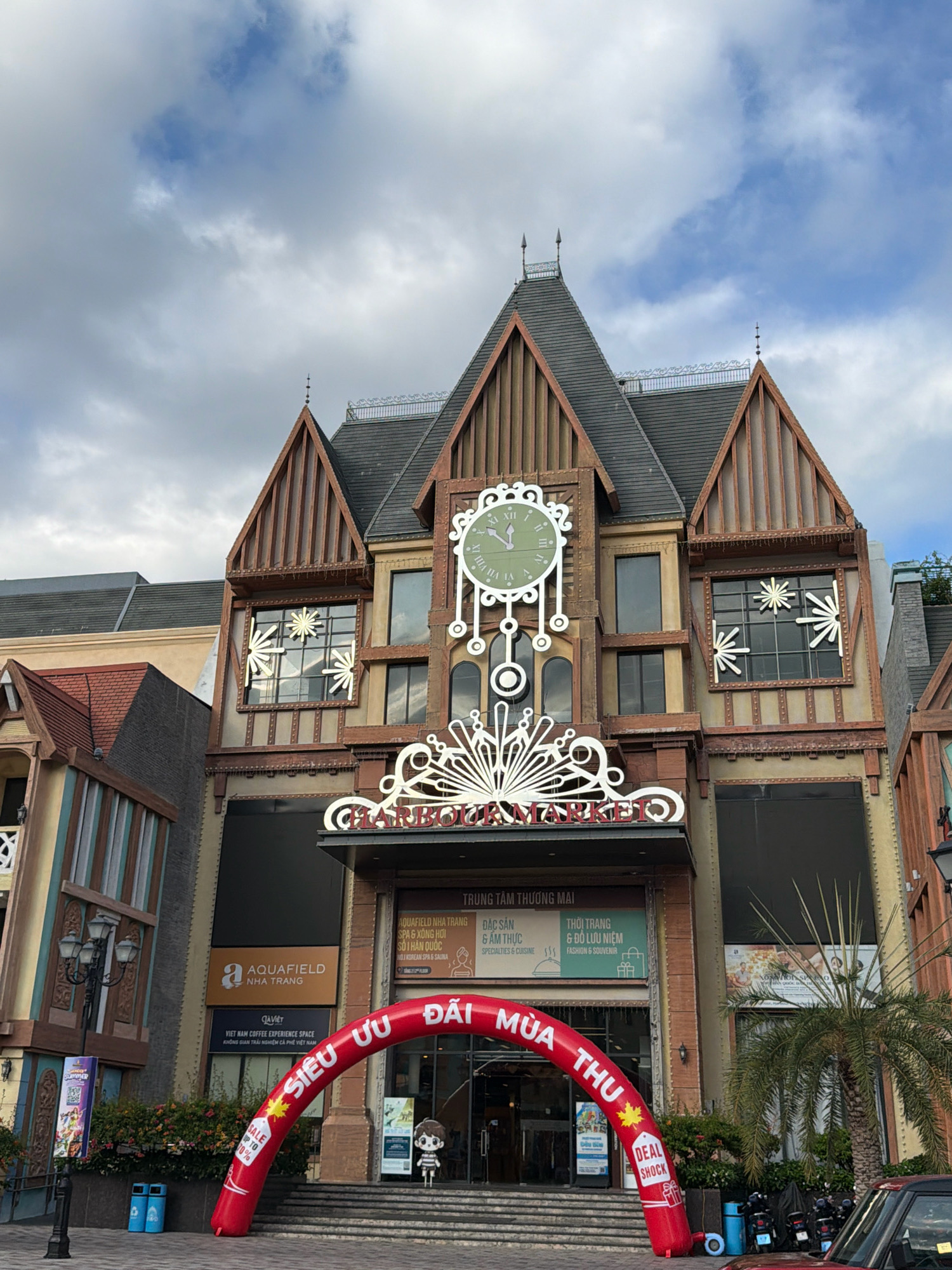 The image shows a wide-angle view of the entrance to a shopping mall in Nha Trang, Vietnam, identified by signage that reads "AQUAFIELD NHA TRANG" and "TRUNG TÂM THƯƠNG MẠI" (Shopping Center). The architecture of the building is elaborate, featuring a large clock on the facade, decorative elements, and a red inflatable archway at the entrance that reads "SIÊU ƯU ĐÃI MÙA THU" (Super Autumn Deals). In front of the entrance, there are stairs leading up to the doors. A small figure dressed in a striped shirt and shorts stands near the entrance. To the left of the entrance, a black lamppost with three lights stands on the sidewalk, and a blue advertising board is visible. Two blue trash cans are positioned near the lamppost. Further to the left, a section of another building with a red tiled roof is visible. To the right of the entrance, a palm tree is situated in front of the building. Several motorcycles are parked to the right of the palm tree, and the rear of a red SUV is visible in the foreground on the right. The sky is partly cloudy.