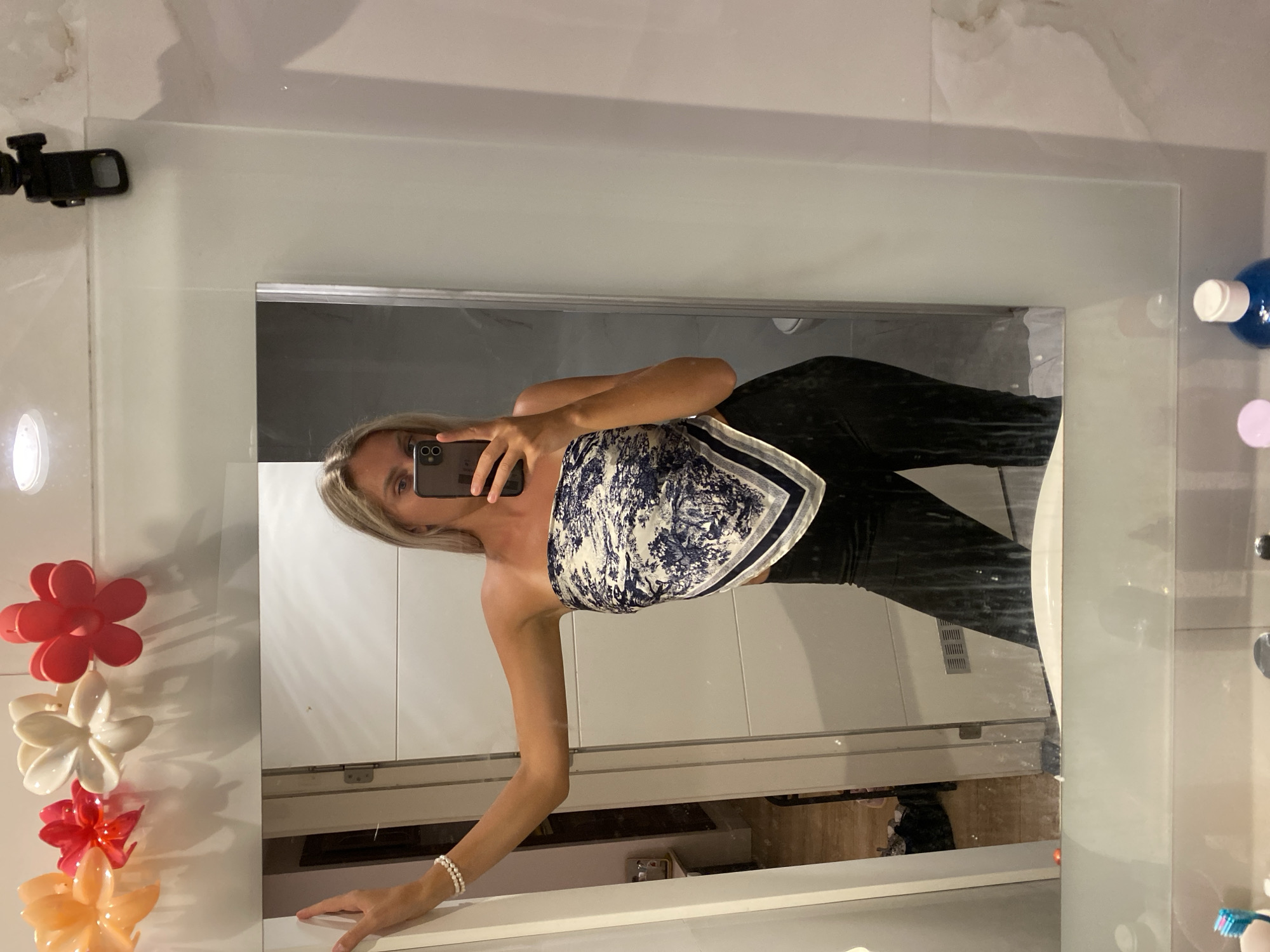 In a bathroom located in Aksu, Turkey, a young woman with blonde hair is taking a mirror selfie. She is wearing a strapless top made from a patterned scarf and black pants. Her left arm rests on the mirror, and she is holding her phone with her right hand to capture the reflection. The mirror reflects a doorway and part of a shower stall. Above the mirror, decorative flower-shaped objects are attached to the wall. The lower part of the mirror shows the bathroom counter with toiletries.