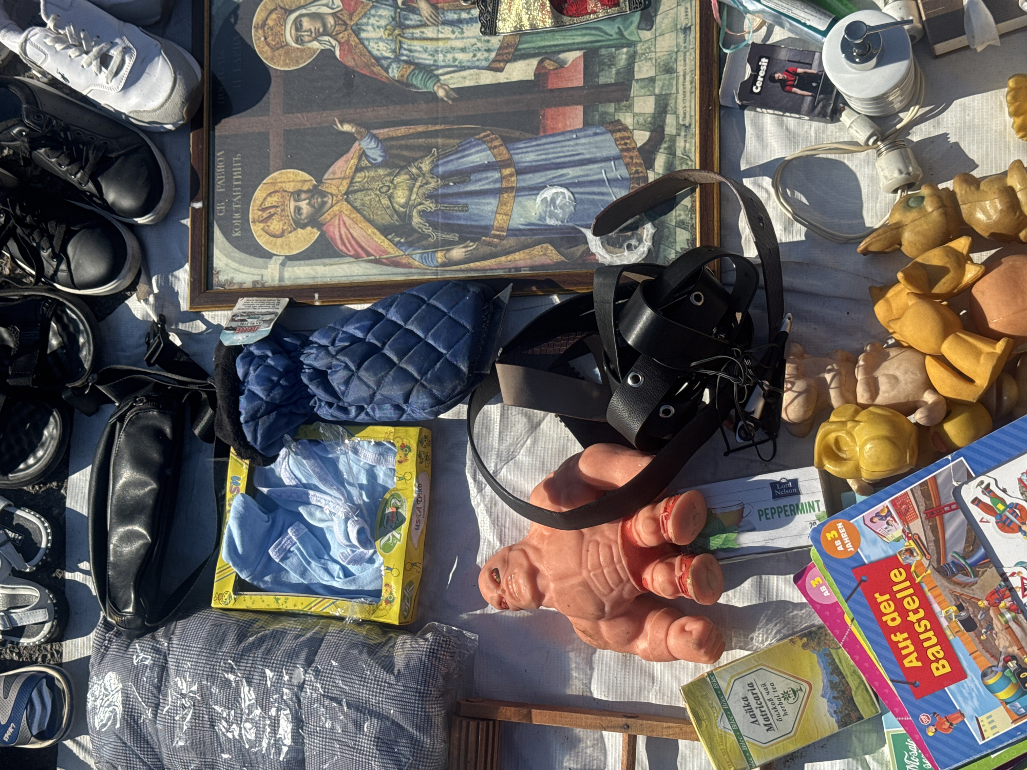 The image depicts an outdoor flea market scene, likely in Sofia, Bulgaria, given the Cyrillic script on some items. The scene is a flat lay of various objects, suggesting a vendor's stall. Prominently featured are several pairs of footwear, including sandals, black lace-up shoes, and white sneakers. There are also clothing items, such as a quilted jacket or blanket in a plastic wrap and a blue quilted pouch.
A key object is a framed religious icon depicting two figures, possibly saints, in traditional attire with halos. Other items include children's books with titles in German ("Auf der Baustelle" and "3 AB JAHREN"), a yellow box containing blue children's clothing, and a toy of a muscular, pink, action-figure-like character. A black leather belt with metal eyelets is also visible, along with packages of tea ("Datika Matricaria herbal tea," "Peppermint").
Smaller items include a "Ceresit" branded product, a lamp fixture with wiring, and several small, yellow, plastic toy figures, resembling animals or cartoon characters. The overall impression is a collection of diverse goods offered for sale in a casual market environment. The ground appears to be covered with a white cloth or tarp.