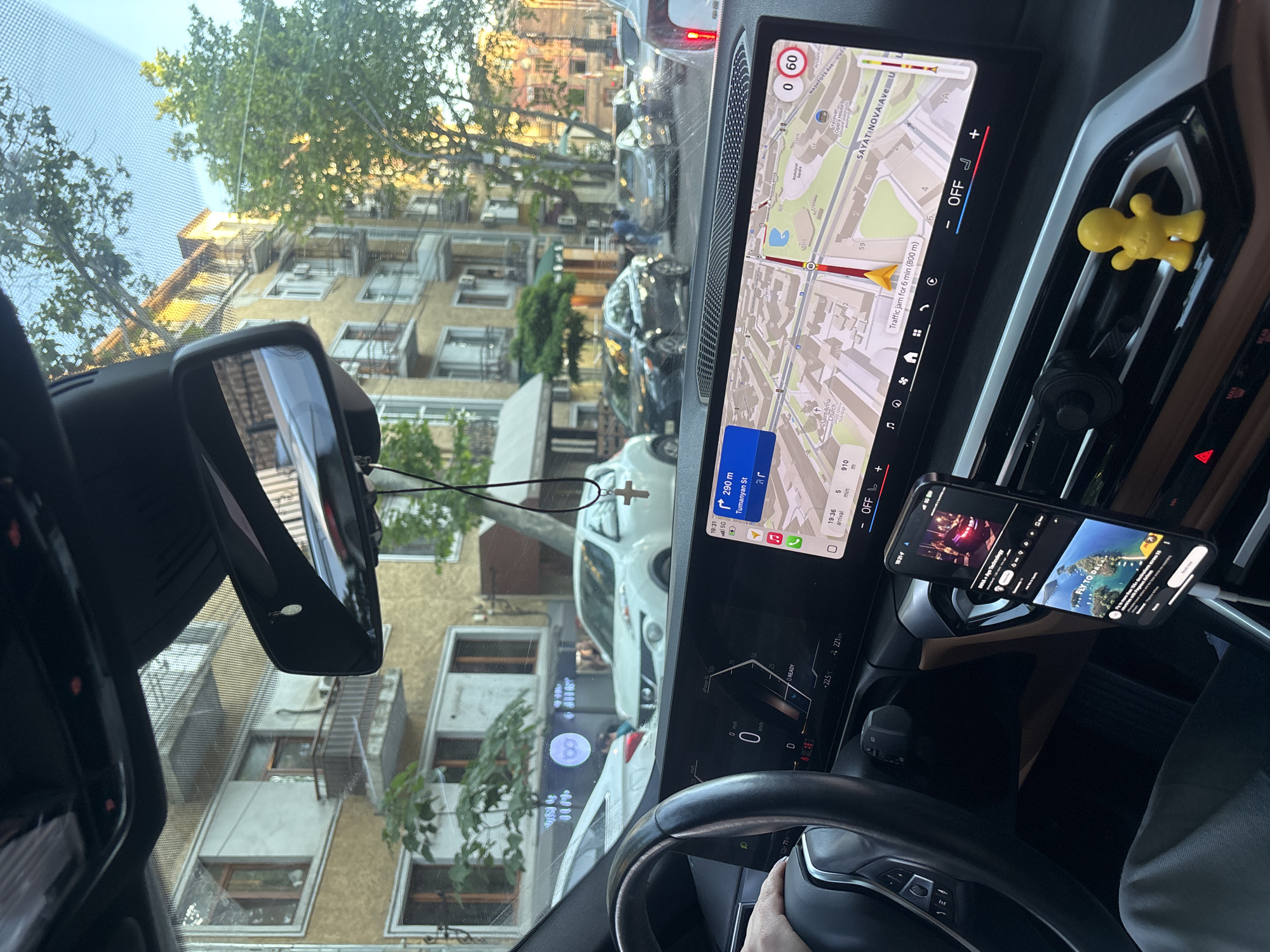 The image captures a view from inside a vehicle, likely a car, as it navigates a street in Yerevan, Armenia. The GPS navigation system on the car's display shows the route, indicating the vehicle is approximately 290 meters from Tumanyan Street. The dashboard also displays a current speed of 0 km/h, with an estimated time of arrival of 19:36 and 5 minutes. The GPS map further shows the traffic jam for 6 minutes (800m).
Through the windshield, one can see the urban landscape of Yerevan, with several apartment buildings visible. The car appears to be stationary in traffic. A small cross hangs from the rearview mirror. A phone is mounted on the right side of the dashboard, displaying content.