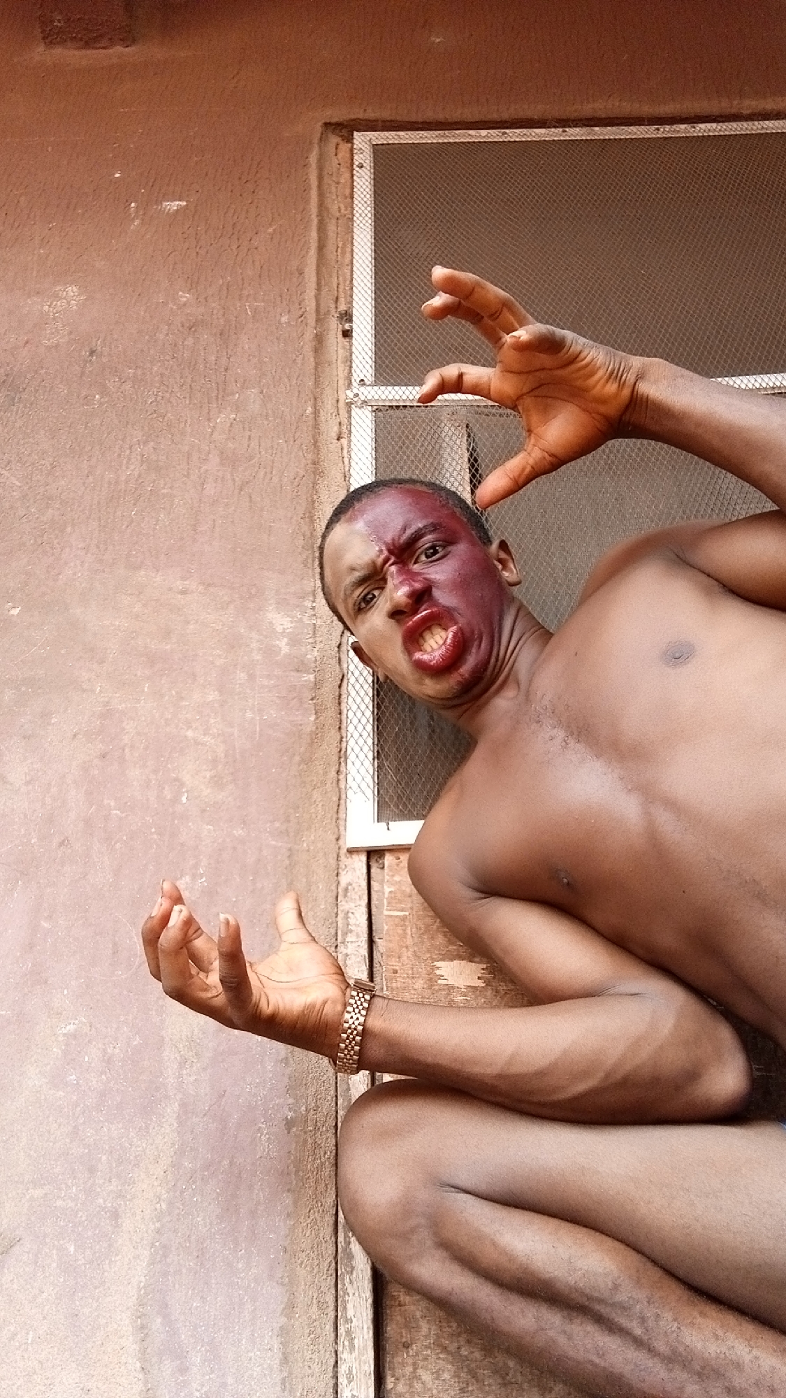 A shirtless dark-skinned man with dark red coloring covering the upper left side of his face poses outdoors, his expression distorted into a grimace, baring his teeth. He holds his hands up with fingers splayed in a clawing gesture. He wears a gold-colored watch on his left wrist. He is positioned in front of a rough, uneven textured wall, with a white-framed window featuring a mesh screen to his right. The location is an unidentifiable outdoor setting in an unknown city.