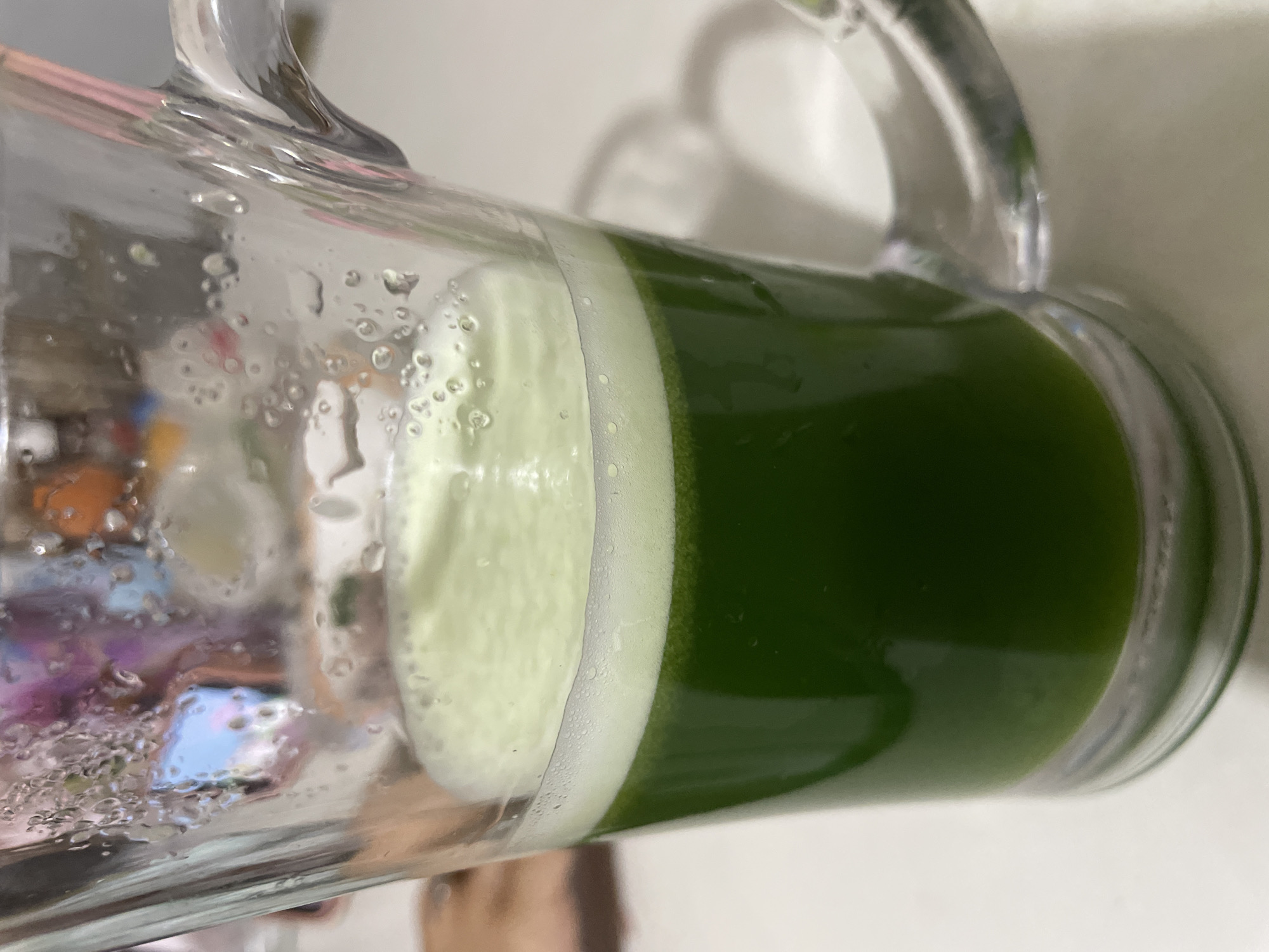 The image shows a close-up of a clear glass mug filled with a vibrant green liquid, topped with a layer of foam. The mug has a handle and appears to be made of glass. Condensation is visible on the exterior of the mug, suggesting it contains a cold beverage. The liquid is a deep, opaque green, with a lighter green, frothy foam on top. The background is blurred and out of focus, but it appears to be an indoor setting with a light-colored surface, possibly a table. Reflections of colorful objects can be seen on the surface of the mug, indicating the presence of other items in the environment. There are no people visible in the image. The image does not provide any specific information or visual cues to determine its location in Davao City, Philippines.
