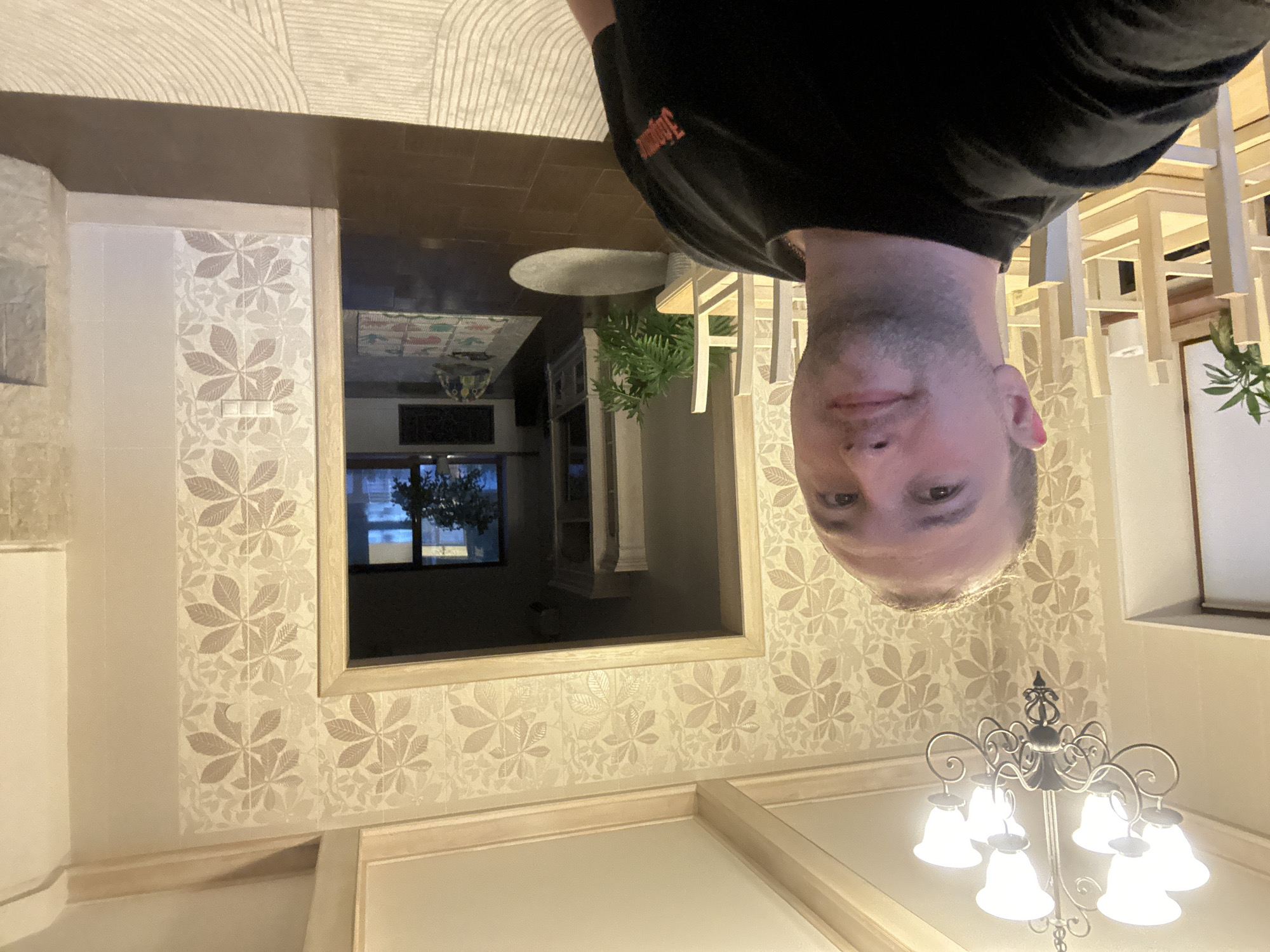 A man in a black shirt with red lettering is positioned upside down in the frame. The interior shows wallpaper with a light-colored floral pattern. In the reflection of what appears to be a doorway or opening, a dining area can be seen with a chandelier and a window. The location is likely an interior of a building in the city of Chaikovsky, Russia, as indicated by the prompt.