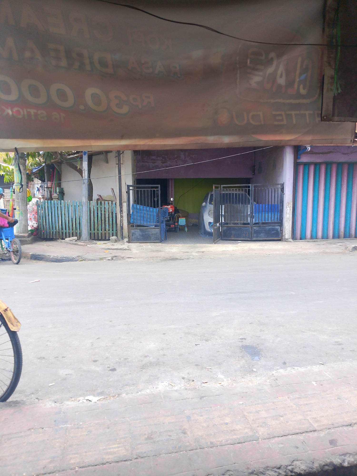 A street scene in Palembang, Indonesia, shows a faded sign with text in English and Indonesian, advertising "Kopi Dream" and " Rp 30.000". The sign is partially obscured by a reflective awning. In the background, a small building with an open garage door is visible, with a white car covered by a tarp inside. A blue and pink striped rolling shutter is on the right side of the building. A green fence is located in front of the building, and a bicycle with a yellow fender is parked on the left side of the image. The ground is paved with concrete and brick.