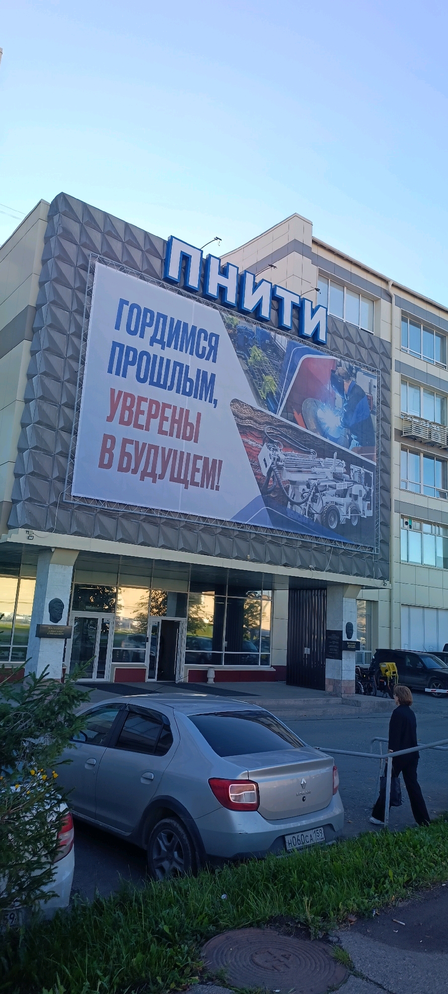 The image shows the facade of the "PNITI" research institute building in Perm, Russia, located at ulitsa Lenina, 86. The building has a large banner on its upper section with blue and red text in Russian that reads "ГОРДИМСЯ ПРОШЛЫМ, УВЕРЕНЫ В БУДУЩЕМ!" which translates to "PROUD OF THE PAST, CONFIDENT IN THE FUTURE!". The banner also features images related to industrial work, including welding and machinery. In front of the building, a silver Renault Logan car is parked, and a woman in dark clothing is walking away from the camera towards the right. The entrance to the building features glass doors and columns. The sky is clear and blue, suggesting it is daytime.