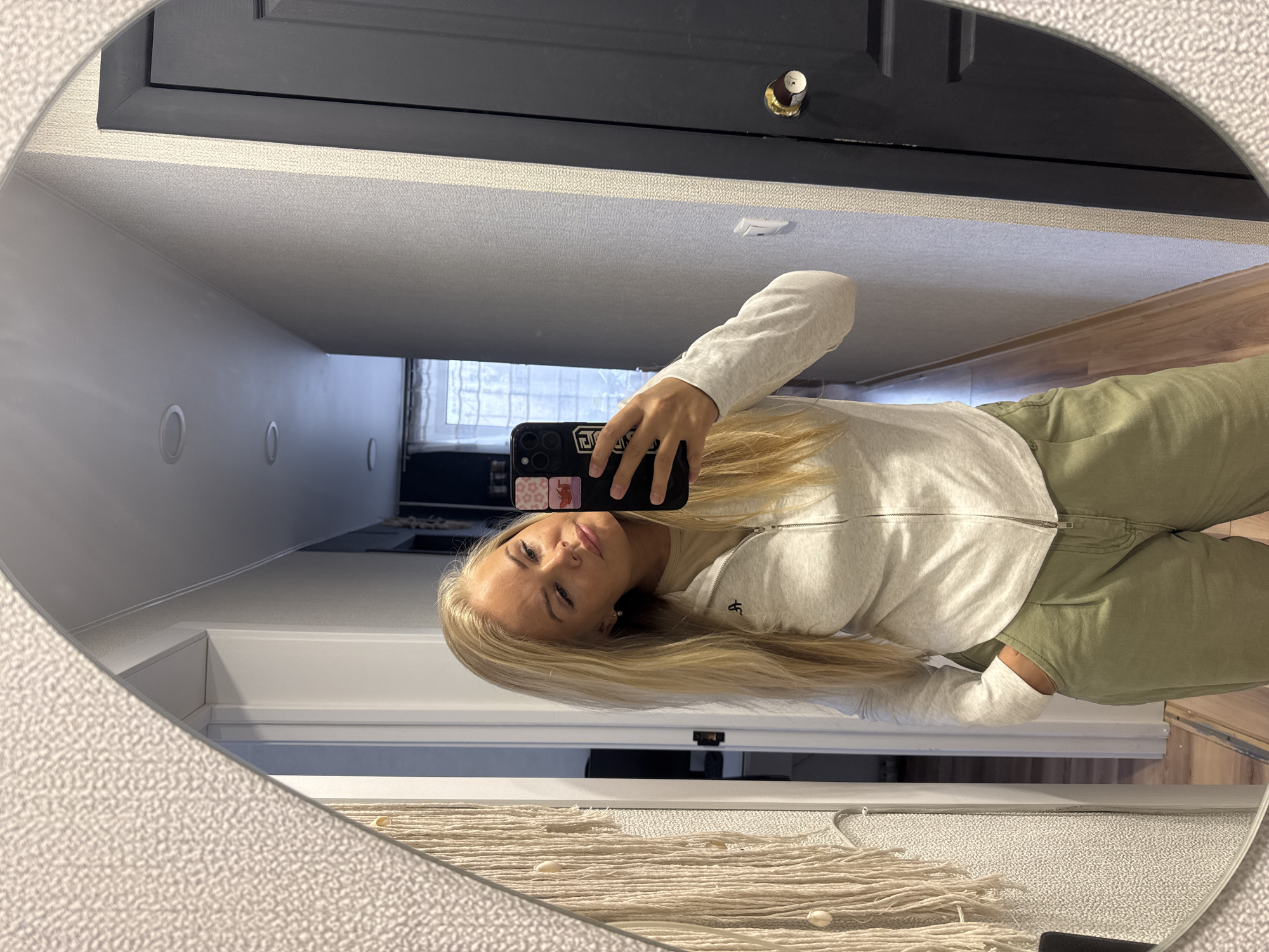 A person is reflected in an oval mirror, holding a smartphone and taking a selfie. The person has long blonde hair and is wearing a light gray zip-up sweatshirt and green pants. The mirror is hanging in a hallway. Behind the person, in the background of the reflection, there is a closed dark-colored door with a brass knob. The hallway has light gray walls with recessed lighting. A macrame wall hanging is visible at the bottom of the mirror. The location is Unknown, Unknown.