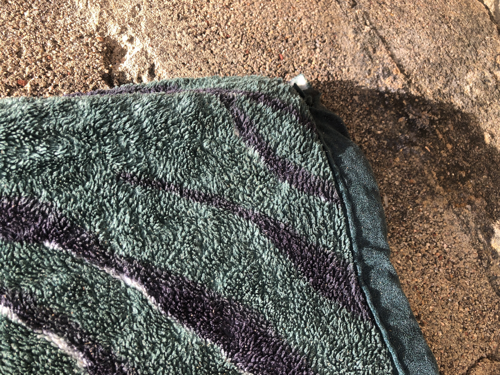 A close-up, overhead shot focuses on a portion of a textured, dark green towel with a dark purple stripe pattern. The towel is draped over a rough, granular surface that appears to be concrete or sand. The lighting suggests it is outdoors, with shadows falling across the scene. The composition emphasizes the textural contrast between the soft fabric of the towel and the coarse texture of the ground.