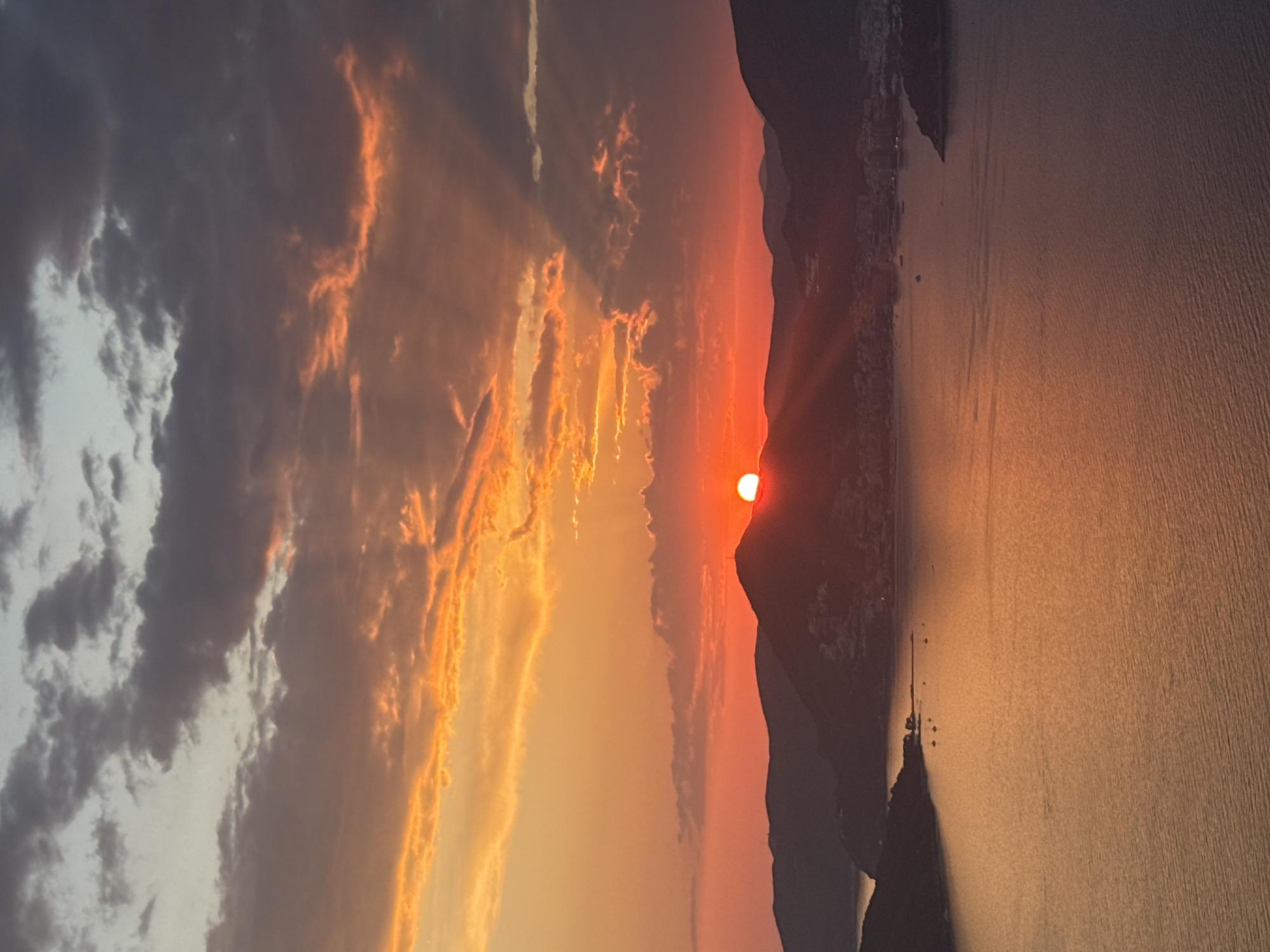 The image depicts a dramatic sunset over a coastal landscape, likely Sveti Stefan, Montenegro, given the context. Silhouetted against the vibrant sky are the distinctive outlines of mountainous terrain and a small, inhabited island connected by a narrow causeway. The sun, a brilliant orange orb, is low on the horizon, casting long rays that pierce through dramatic clouds, painting them in hues of orange and gold.
The sea stretches out, reflecting the warm colors of the sunset, creating a shimmering, almost ethereal surface. The texture of the water suggests a calm or gently rippling state. Along the coastline, some structures are visible, hinting at human presence, perhaps a village or resort. The overall mood is serene and awe-inspiring, with the powerful natural light dominating the scene. The time of day is clearly sunset, marked by the low position of the sun and the intense coloration of the sky and its reflections. The weather appears clear with scattered clouds that are illuminated by the setting sun. There is no visible text in the image.