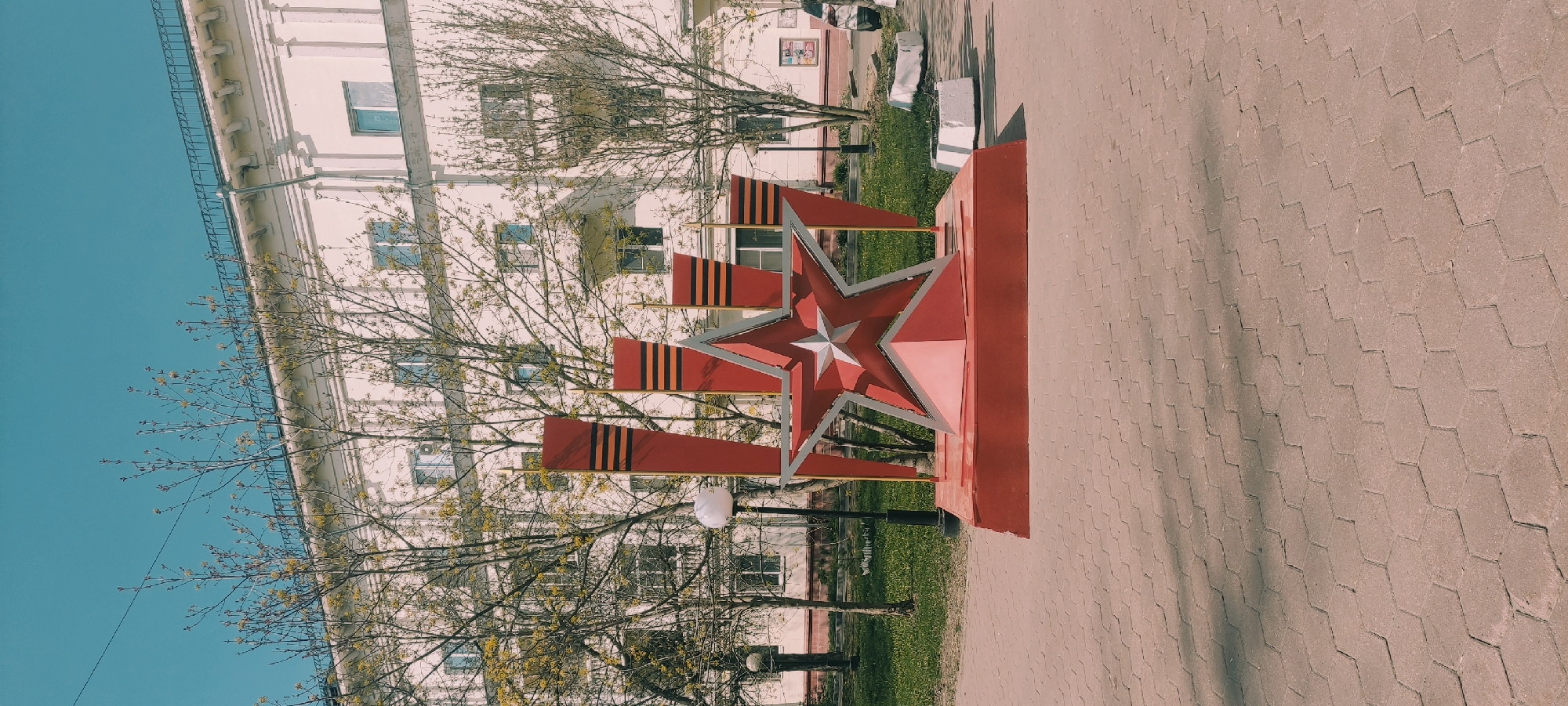 The image captures a red star monument, presumably a memorial or a symbolic structure, situated in Fryazino, Russia. The star is prominently displayed on a red base, accompanied by what appear to be red banners adorned with black and orange stripes. These elements are positioned on a paved area, likely a sidewalk or a square, adjacent to a patch of grass. The background includes a white multi-story building with windows and a few trees. The sky is visible, a clear and light blue. The photograph is taken outdoors during daylight hours.