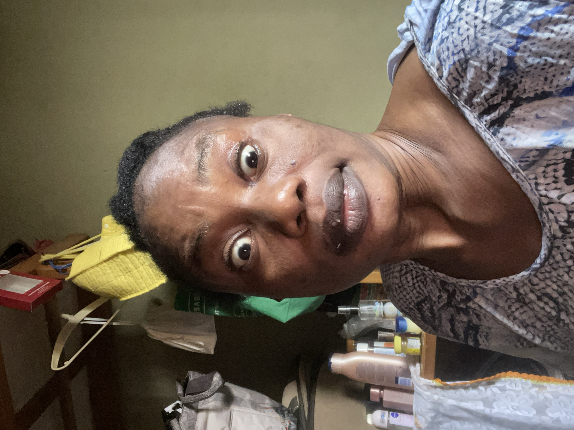 The image features a close-up of a Black woman, viewed from a slightly low angle, looking directly into the camera. She has short, dark, textured hair and a wide-eyed expression with slightly pursed lips, suggesting surprise or curiosity. Her skin appears moisturized, and some minor blemishes are visible on her forehead. She is wearing a top with a patterned design in shades of blue, grey, and white, possibly depicting a snakeskin or abstract motif.
The background indicates an indoor setting in Narayi, Nigeria, with various household items on shelves or tables. To her left (when the image is viewed upright), a bright yellow textured fabric item, a partially visible red box, and a white clothes hanger are present. Below her chin, a green fabric item, possibly a face mask, is discernible. To her right, a collection of bottles, likely toiletries or cosmetics, are lined up. These bottles vary in color, including light brown, white, and clear, with different colored caps. A label on one bottle appears to partially read "NIVEA".
The lighting suggests an indoor environment, possibly with artificial light or indirect daylight. The overall impression is a candid moment, likely a selfie, within a personal space that appears somewhat lived-in. No specific activity other than posing for the photograph is depicted. Visible text includes "NIVEA" on a bottle and partially obscured text on a green item below her chin.