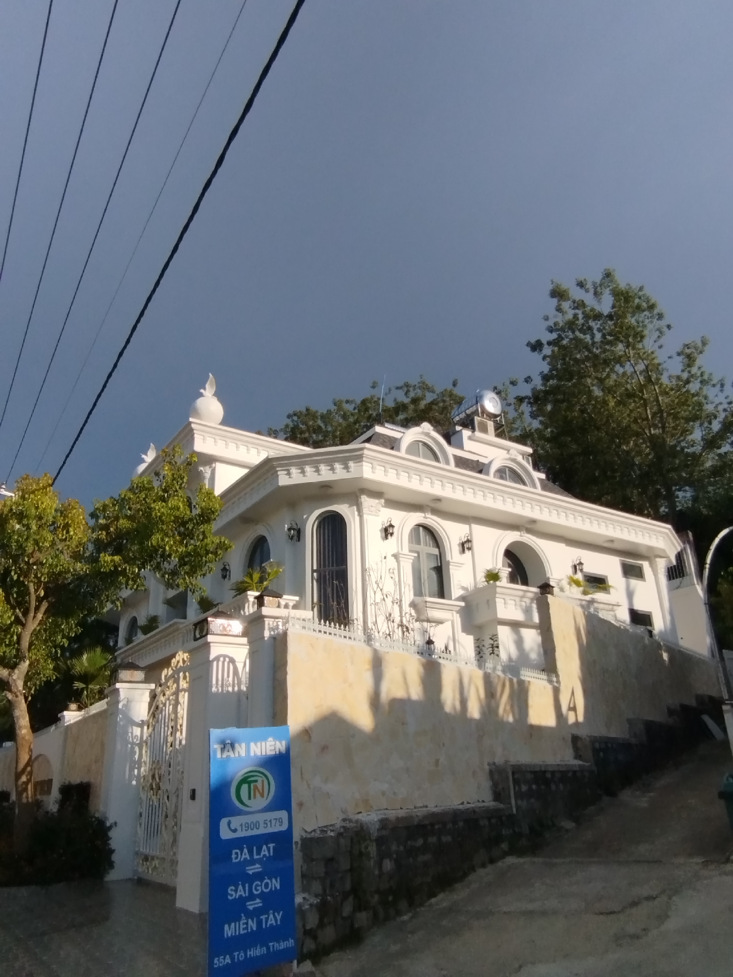 The image shows a white building with ornate architectural details, including arched windows, decorative trim, and a rounded dome topped with a bird sculpture. The building appears to be a residence or a business, with a large white metal gate in front. A blue sign with Vietnamese text and a phone number is placed near the gate. The building is situated on a sloped terrain, with a stone wall and a pathway leading up to it. Trees surround the building, and power lines are visible in the upper left corner of the image. The sky is clear and blue. Based on the text on the sign, which includes "ĐÀ LẠT" and "SÀI GÒN", the location is likely in or near Da Lat, Vietnam. The text "55A Tô Hiến Thành" suggests an address.