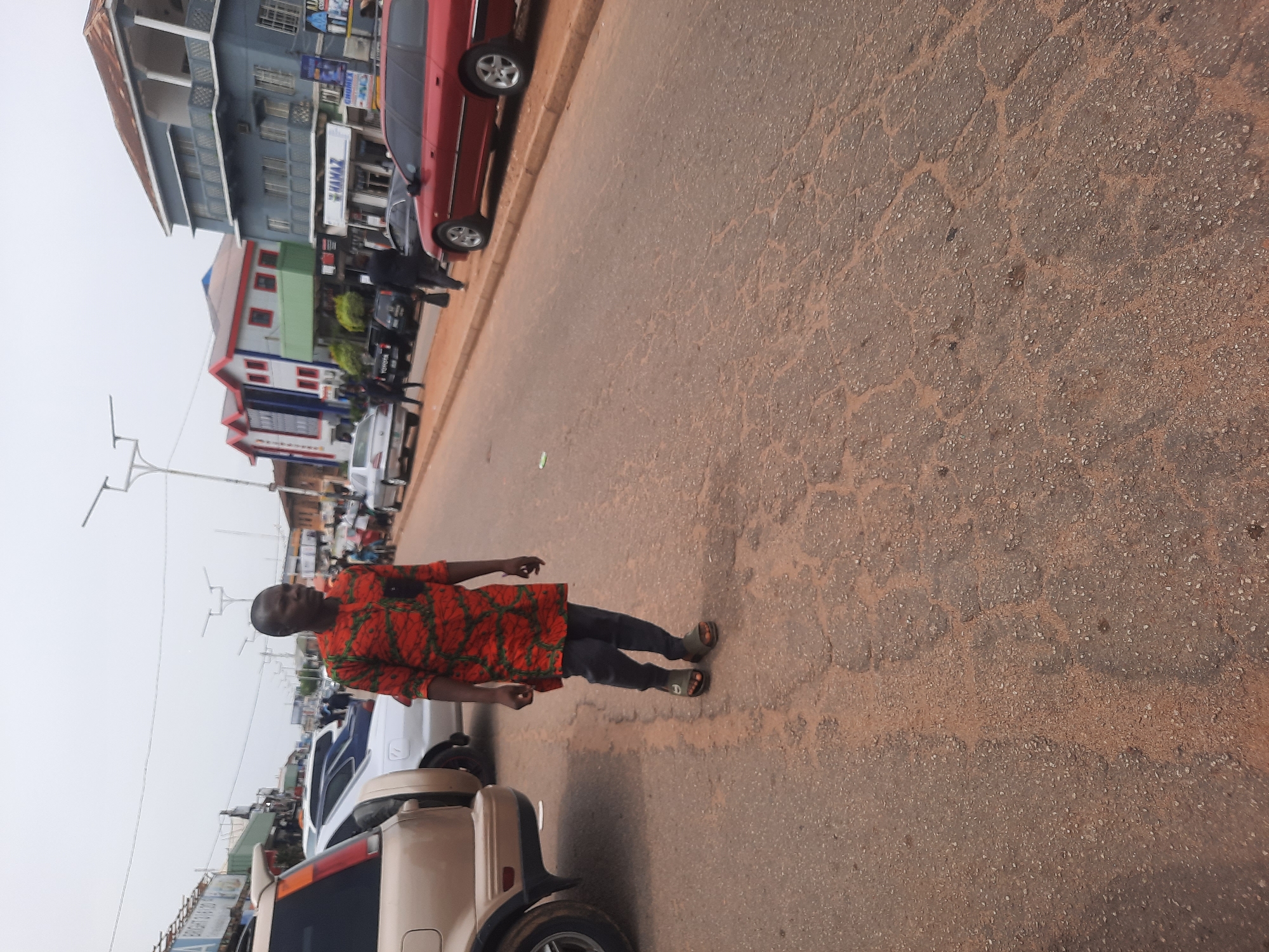 The image shows a man walking on a paved road with cars parked along the side in Jos, Nigeria. The road surface is cracked and uneven. The man is wearing an orange patterned shirt, dark pants, and sandals. Buildings are visible in the background, with visible commercial signage. Several parked cars are along the left side of the road. The sky appears overcast. Streetlights are visible in the distance.