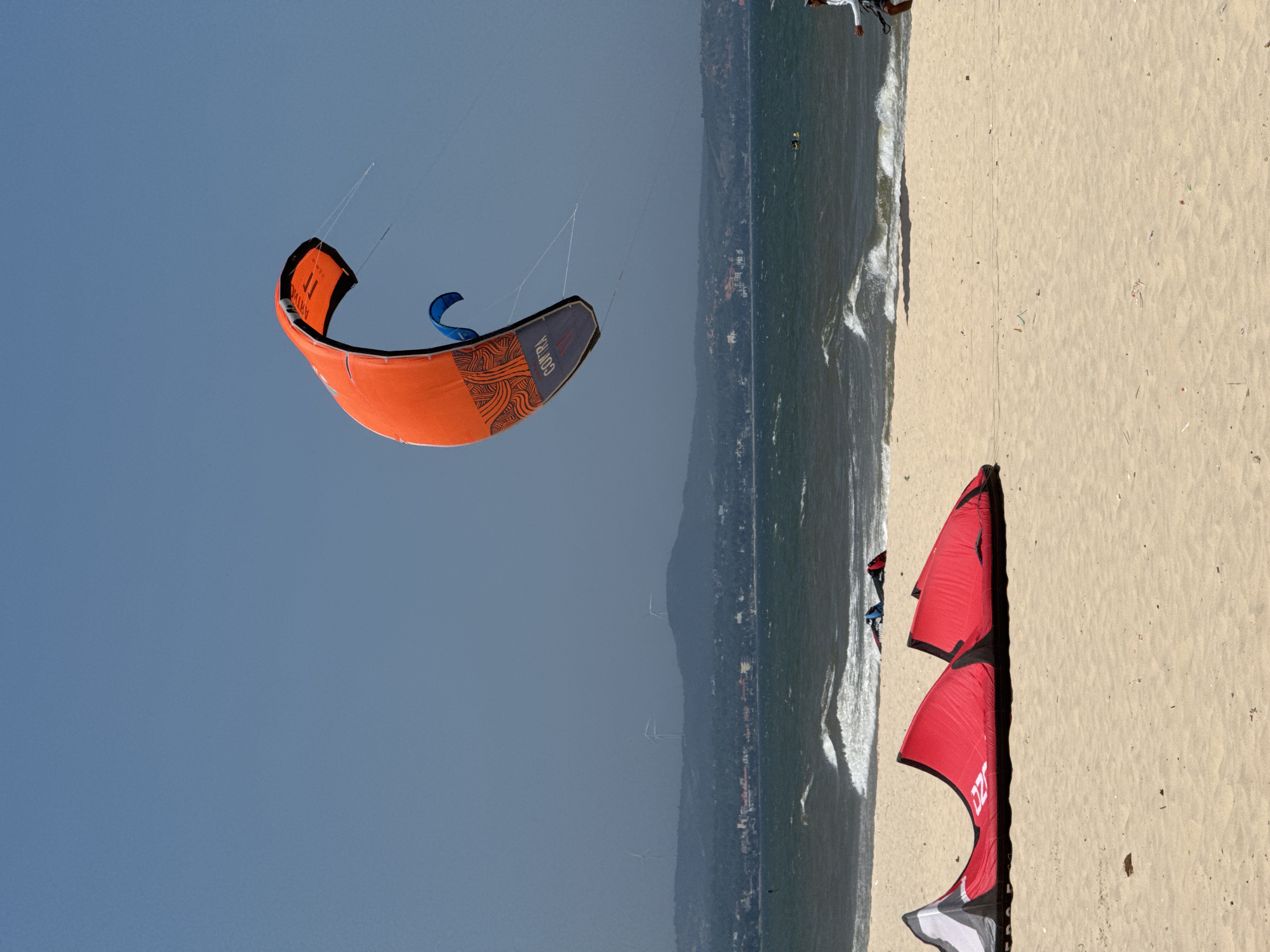 This is a photograph taken on a sunny day in Mũi Né, Vietnam. The image depicts a beach scene with kitesurfing activities in progress. In the foreground, there are two red kites lying on the sandy beach. In the middle ground, the ocean has moderate waves. Further out in the water, two people can be seen kitesurfing. In the background, there is a coastline with buildings and hills, and wind turbines are visible on the hills. The sky is clear and blue.