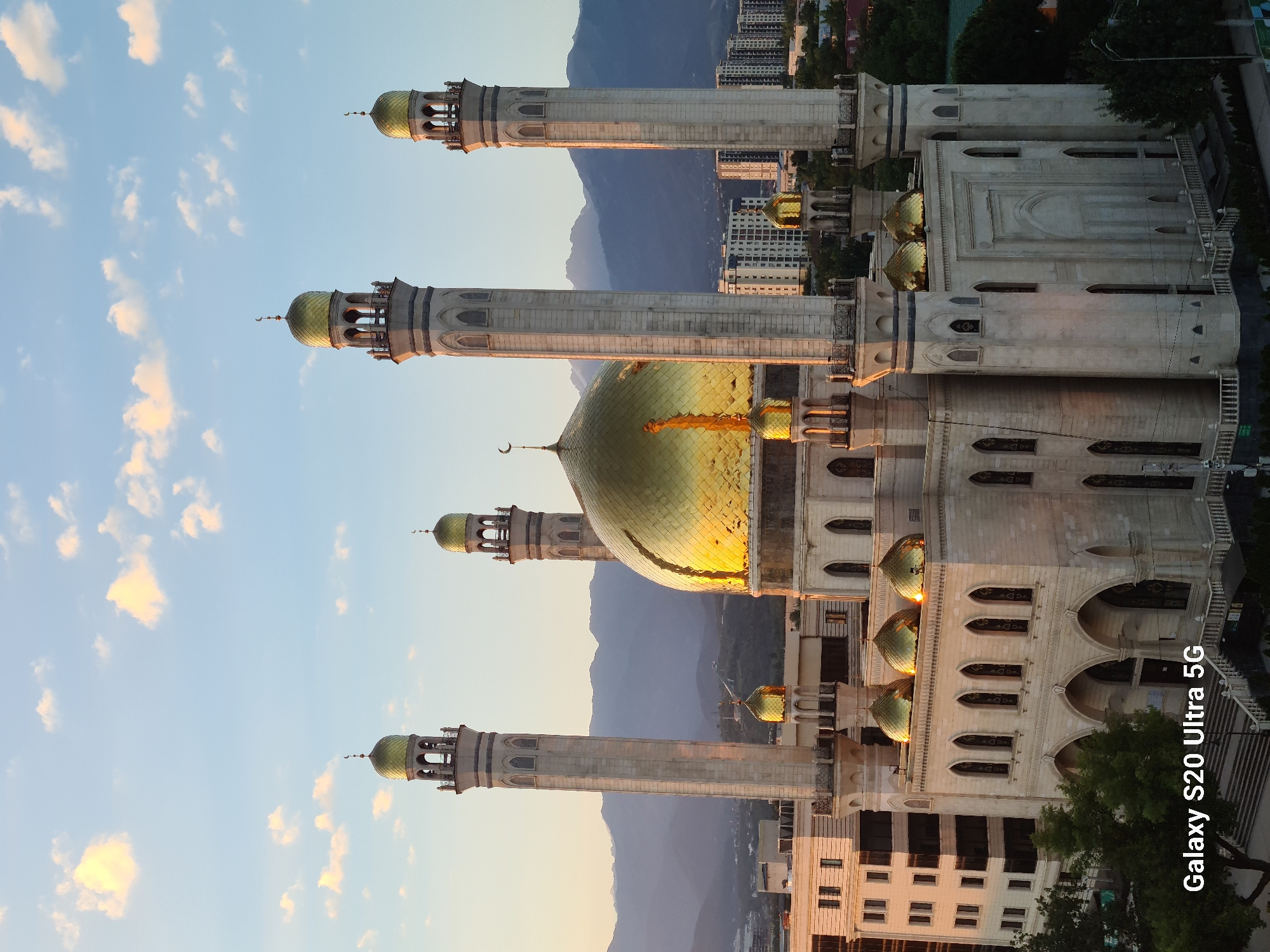 The image shows a large, ornate mosque with multiple gold-domed minarets and a central golden dome. The mosque is constructed of light-colored stone and features intricate architectural details, including arched windows and balconies. The mosque is situated in an urban environment, with modern buildings and apartment complexes visible in the background. Distant mountains form a backdrop to the city. The sky is clear with a few scattered clouds, and the lighting suggests it is either sunrise or sunset. The text "Galaxy S20 Ultra 5G" is visible in the lower left corner, indicating the device used to capture the photograph. This is the Central Mosque of Almaty, Kazakhstan.