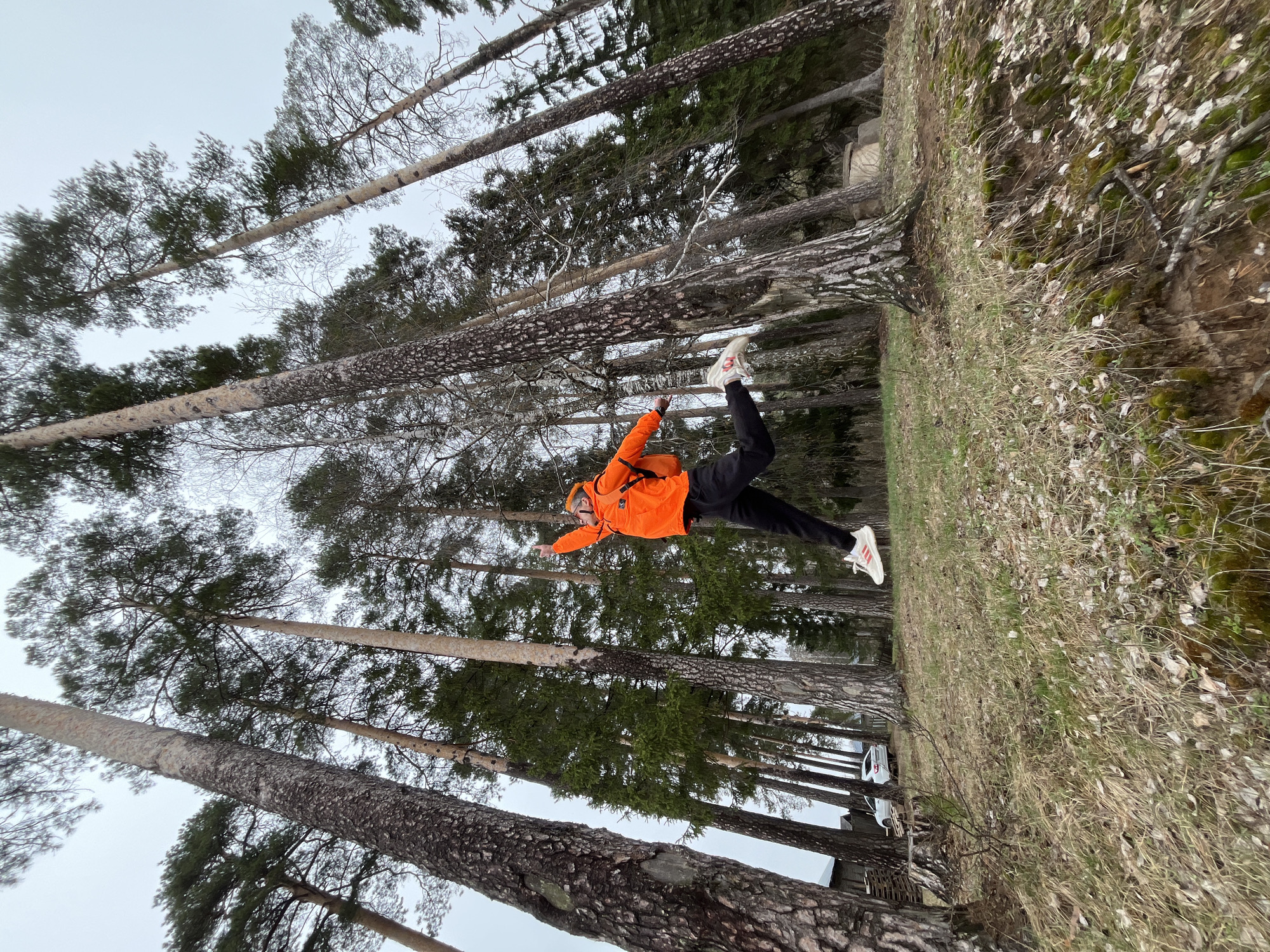A person is captured mid-air in a forest in Chaikovsky, Russia. The individual is wearing an orange jacket and black pants with white sneakers. The pose suggests movement or a jump. Tall trees surround the subject, and the ground is covered with vegetation.