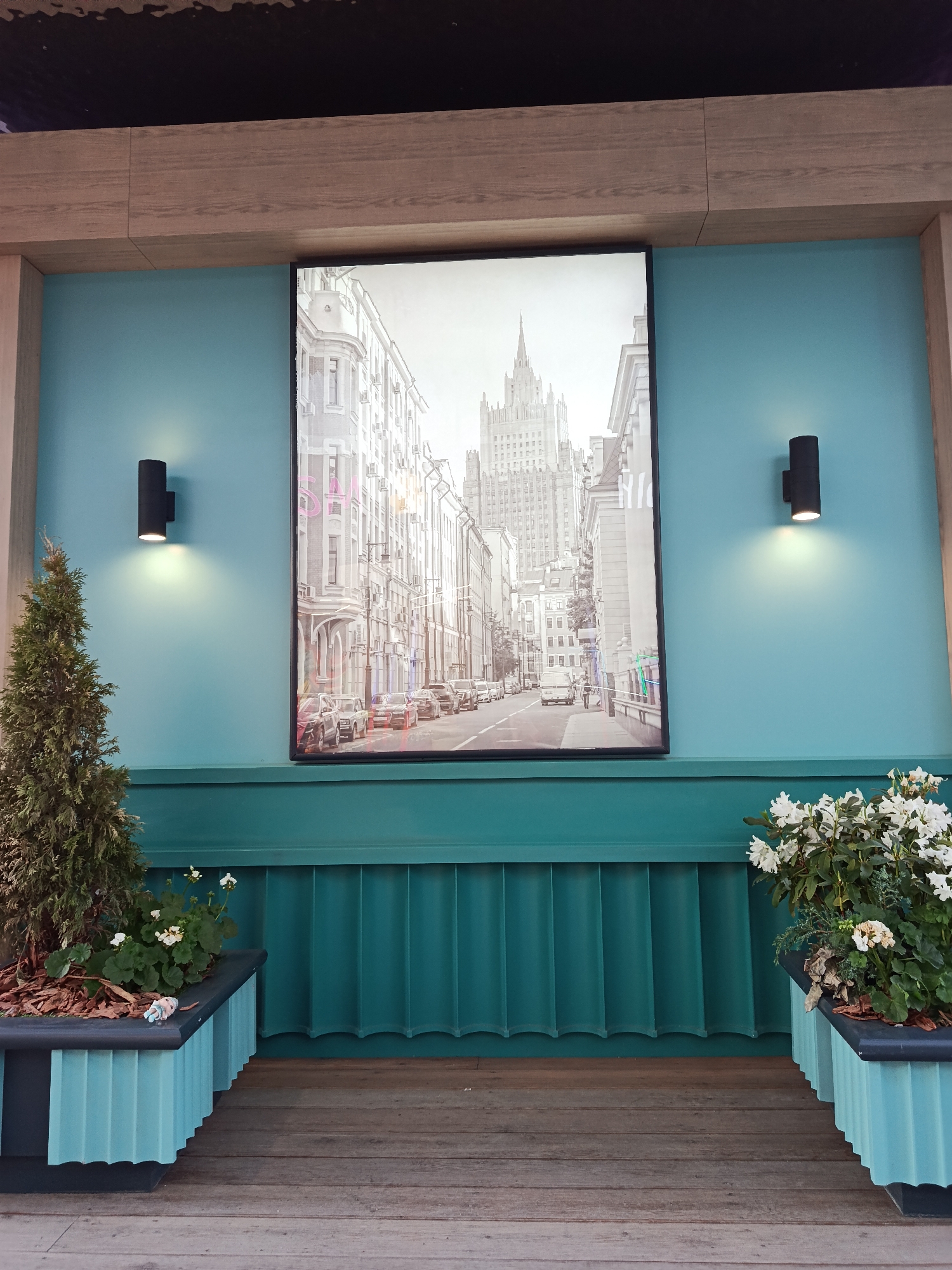 The scene presents a staged, indoor setting reminiscent of a public space in Moscow, Russia. A framed black-and-white photograph dominates the center of the teal-colored wall, depicting a street scene with the Seven Sisters skyscraper, specifically the Ministry of Foreign Affairs, prominently featured in the background.
Flanking the photograph are two cylindrical black wall sconces, each casting a warm light that highlights the smooth teal wall surface. Below the photograph, a teal decorative wainscoting runs horizontally, providing a visual break between the wall and the floor.
In the foreground, two identical planters sit on a wooden planked floor. The planters are teal with a dark gray top, and feature fluted sides. The left planter contains a small coniferous tree, accompanied by smaller flowering plants with white blossoms, set in a bed of wood chips. The right planter mirrors this arrangement, featuring flowering plants with white blossoms. The entire setting appears to be designed to evoke a sense of place, specifically referencing the urban landscape of Moscow through the photograph and color scheme.
