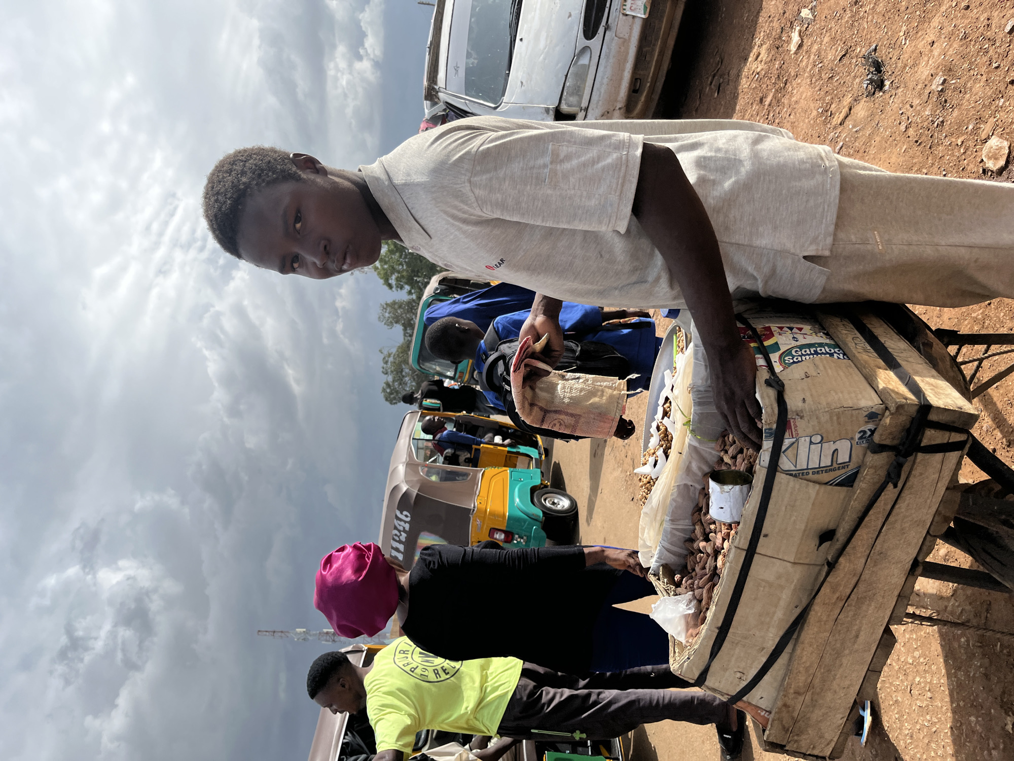In Rafin Par, Nigeria, under a cloudy sky, a young male vendor stands beside a cart laden with peanuts. He wears a light beige polo shirt and matching sweatpants. The cart, constructed from wood and cardboard, displays peanuts in plastic bags and loose, alongside a discarded can. In the background, various vehicles, including a yellow and green auto-rickshaw and a white car, are present, suggesting a roadside market or transportation hub. Other individuals, including a person in a bright yellow shirt and another wearing a pink head covering, are also visible, engaging in activities related to the vehicles and market. The ground is composed of reddish dirt.