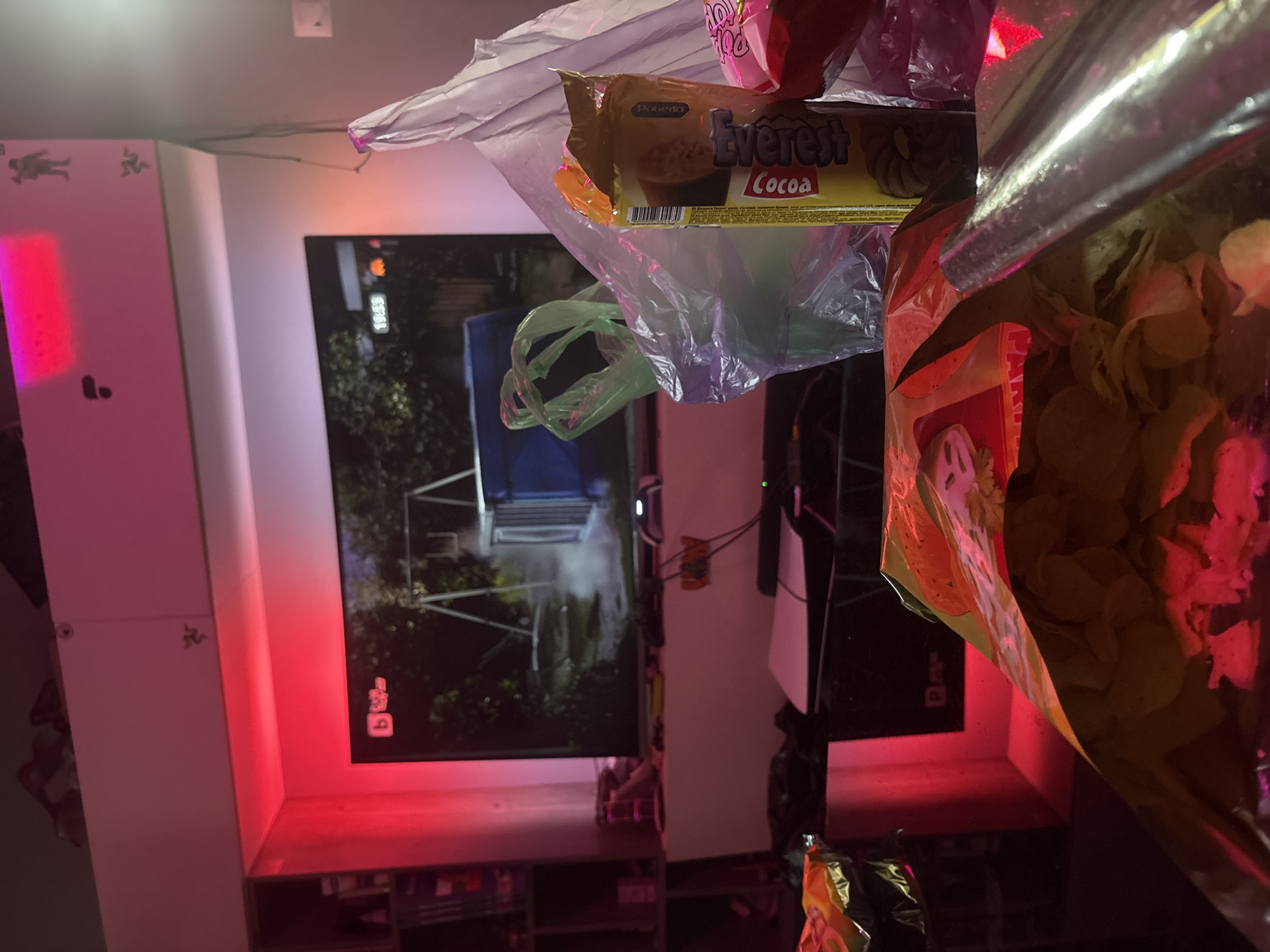 This image depicts an indoor scene in Sofia, Bulgaria, characterized by low light conditions and a dominant red ambient glow from LED strip lighting. The perspective is from a low angle, looking towards a wall-mounted television and an entertainment setup.
In the immediate foreground, several open snack packages and plastic bags are visible on a surface, likely a table. This includes an open bag of light-colored potato chips, another partially visible bag of snacks with orange/yellow packaging, and a standing yellow package of "Everest Cocoa" branded biscuits with "Dobrada" also visible. Multiple clear, green, and purple translucent plastic bags are distributed among these items.
In the midground, a white entertainment unit or shelf is situated beneath the television. On this unit, a white PlayStation 5 console is prominent on the left. Various cables, a black gaming controller (partially obscured), and other electronic devices are present on the surface.
The central focus is a large flat-screen television displaying an outdoor scene. The screen shows a green environment with trees, a metal swing set, and a blue container or dumpster on a dirt path. In the top-left corner of the screen, a "b" logo and "sky" are visible, alongside a digital clock displaying "19:53" in the top-right.
Above and to the left of the television, open shelves contain various items, including books. Above the television, white overhead cabinets are mounted. On the right side of these cabinets, a distinct Razer logo sticker (three-headed snake) is affixed, with another similar logo slightly higher. Small, dark figurines or decorations are also placed on the top surface of these cabinets. The wall on the far right features a light switch or outlet. No individuals are visible in the scene.