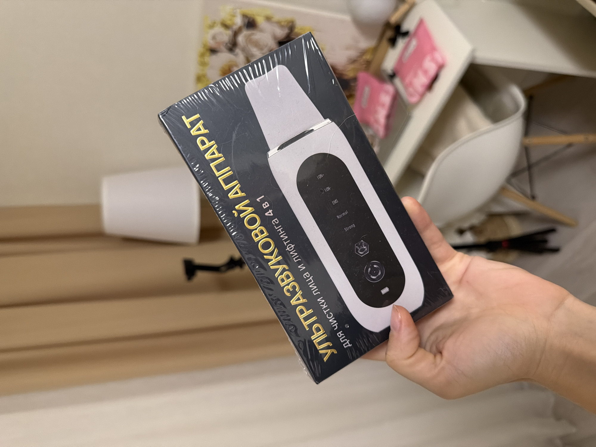 A person's hand holds a black and silver box containing a white ultrasonic skin scrubber. The box has text in Russian that translates to "ULTRASONIC APPARATUS for facial cleansing and lifting 4 in 1". The scrubber itself is visible, with buttons labeled "ION+", "ION-", "EMS", "Normal", and "Strong". The background is out of focus but appears to be an indoor setting with shelves, a white chair, and what looks like a desk or table. The lighting suggests it could be daytime.