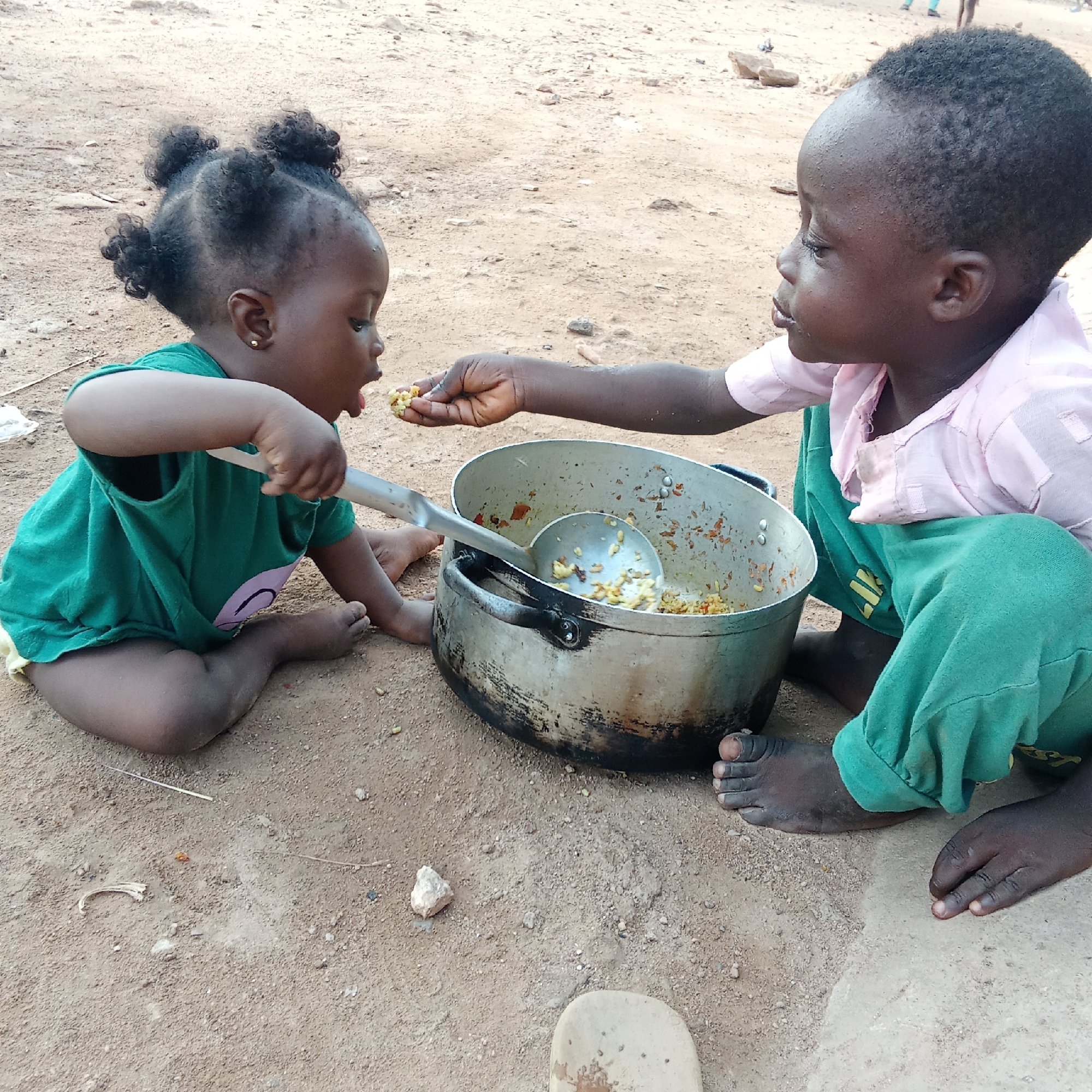 Two young children are sharing a meal directly from a large, well-used aluminum cooking pot, seated on the bare, sandy ground. The scene appears to be outdoors in a dry, warm environment in Akwanga, Nigeria, indicated by the natural light and dusty earth.
On the left, a younger child, likely a toddler, has dark skin and curly dark hair styled in small puffs, adorned with tiny gold stud earrings. This child wears a green t-shirt and light-colored undergarment, and is shown with an open mouth, eagerly accepting food. Their right hand loosely holds the handle of a large metal serving spoon that rests inside the pot.
On the right, an older child, also with dark skin and short, dark, curly hair, is dressed in a light pink collared shirt over a green garment. This child attentively offers a small portion of the meal from their hand to the younger child's mouth, demonstrating a moment of care and sharing.
The pot contains a visible amount of cooked food, appearing to be grains like rice, mixed with small pieces of vegetables or spices. The ground is a light brown, sandy texture, with scattered small rocks and debris. A light-colored object, possibly a piece of footwear, is partially visible in the bottom right corner. The overall atmosphere conveys a simple, everyday moment of nourishment and sibling interaction.