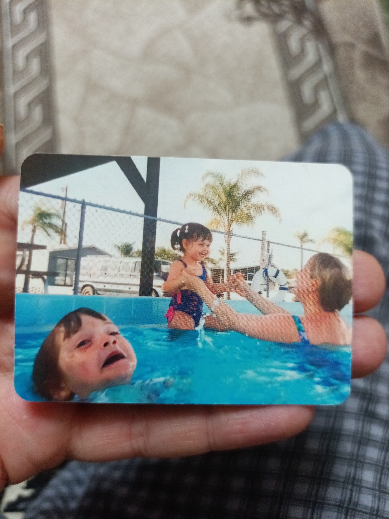 The image is a photograph of a photograph, held by a person's hands. The inner photograph depicts three individuals in a bright blue swimming pool. In the foreground, to the left, a young child with brown hair is partially submerged, with their head and shoulders visible above the water. The child's eyes are wide open, and their mouth is open, appearing to be crying or shouting. To the right, a young girl with dark hair tied in pigtails is in the pool, wearing a blue and pink swimsuit. She is holding hands with an adult woman who is facing away from the camera, also in the pool and wearing a blue swimsuit. In the background, there is a chain-link fence, some palm trees, and what appears to be a building or RV. The scene is outdoors, suggesting a recreational area.
The image does not provide any information or visual cues to indicate it is located in the city of Nyagan, Russia.