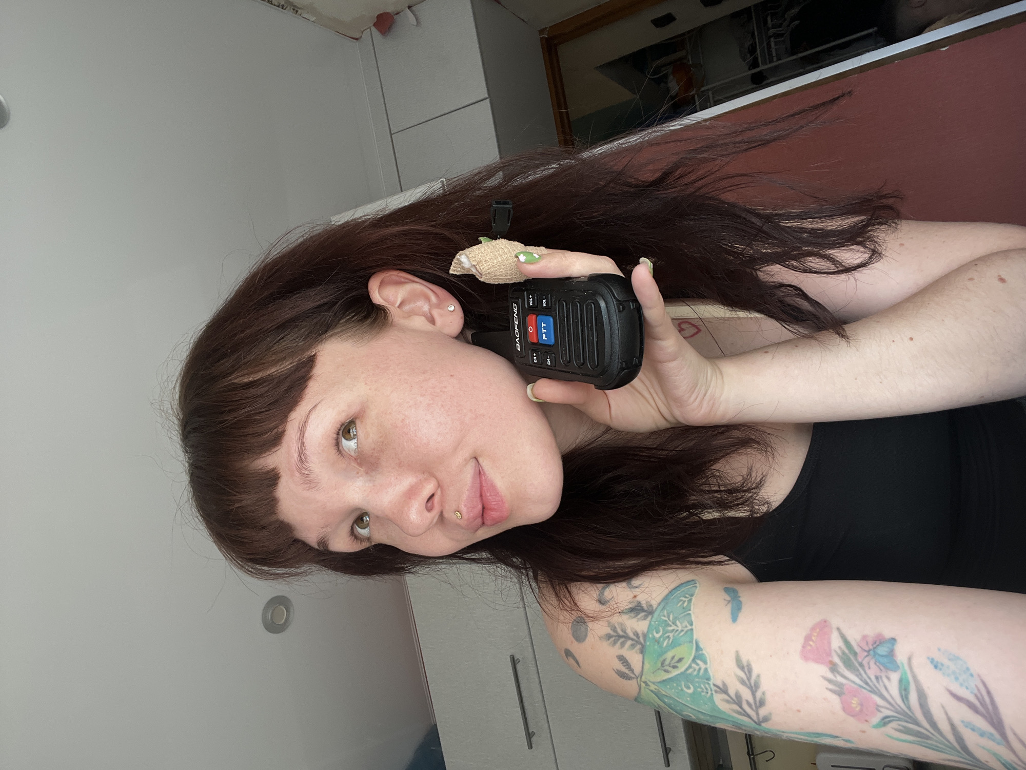 The media file depicts a young person with long, dark brown hair and fair skin, holding a black Baofeng walkie-talkie to their ear. The person is looking towards the camera with a slight smile and appears to have light-colored eyes. They are wearing a black tank top, and their left arm, visible in the lower right of the frame, has tattoos of various plants and insects in shades of green, blue, and pink. There is also a small silver stud piercing on the person's philtrum. A bandage is wrapped around the person's index finger, which is pressing the "PTT" button on the walkie-talkie. The background appears to be an indoor setting with a light-colored wall and cabinets. The orientation of the image suggests it was taken in a personal environment, possibly a home or dressing room. There is no clear indication of the time of day or weather. The person's expression conveys a sense of engagement and perhaps amusement. The walkie-talkie has visible labels for "VOL+", "VOL-", "CH+", "CH-", and "PTT" in red and blue. The brand "BAOFENG" is clearly visible on the device.