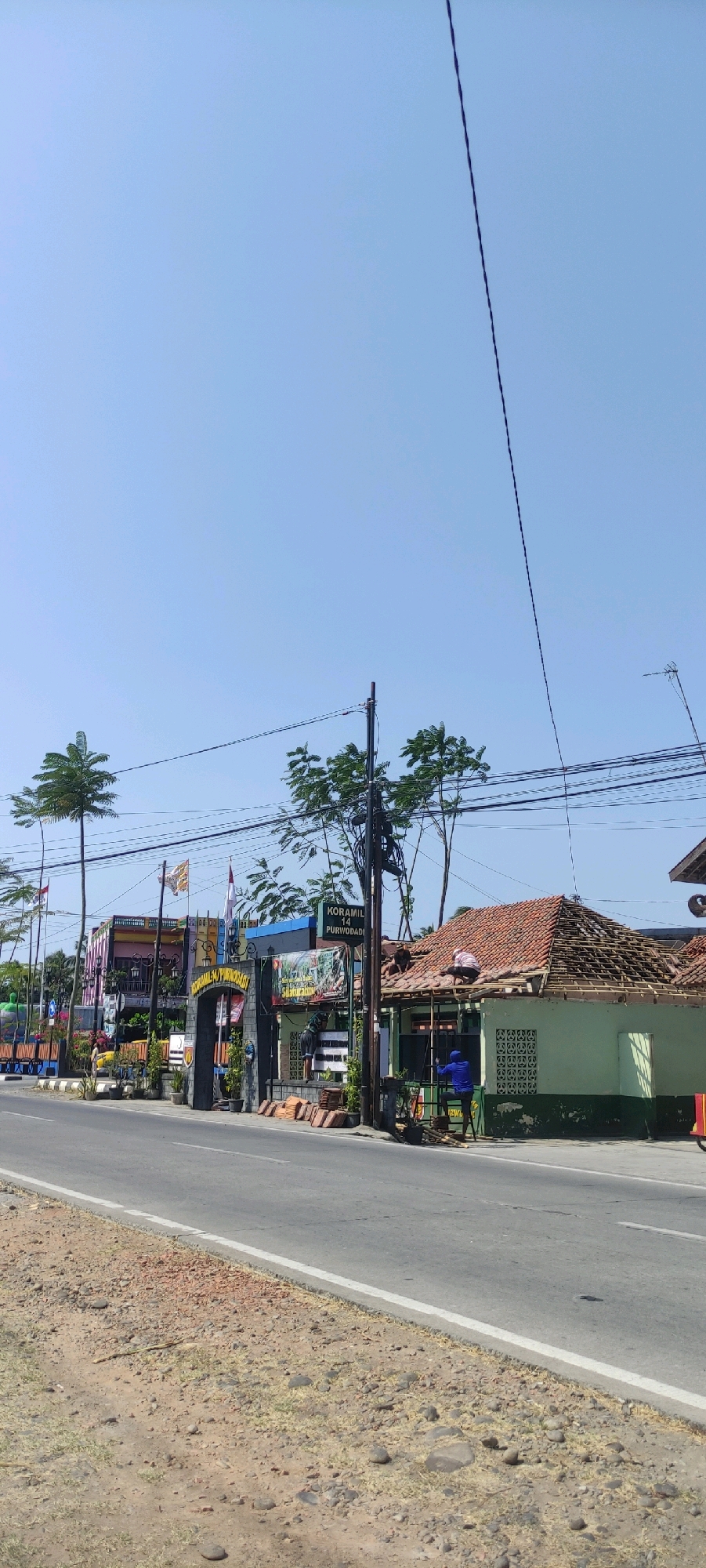 The image depicts a sunny daytime scene on a roadside in Purwodadi, Indonesia, likely near the Koramil 14 Purwodadi. The foreground shows a dusty shoulder and asphalt road with a white line marking the edge. In the midground, an arched gateway with signage reads "KORAMIL 14 PURWODADI". To the right of the gateway, a light green building with a red tile roof is undergoing repairs; several individuals are visible on the roof working, and one person in a blue shirt is standing on a ladder. Various building materials, including stacks of red roof tiles, are scattered around the construction site. Behind the gateway and to the left, a colorful building complex with flags and banners is visible. Several palm trees and other foliage are interspersed throughout the scene, and electrical wires and poles are prominent. The sky is clear and blue.