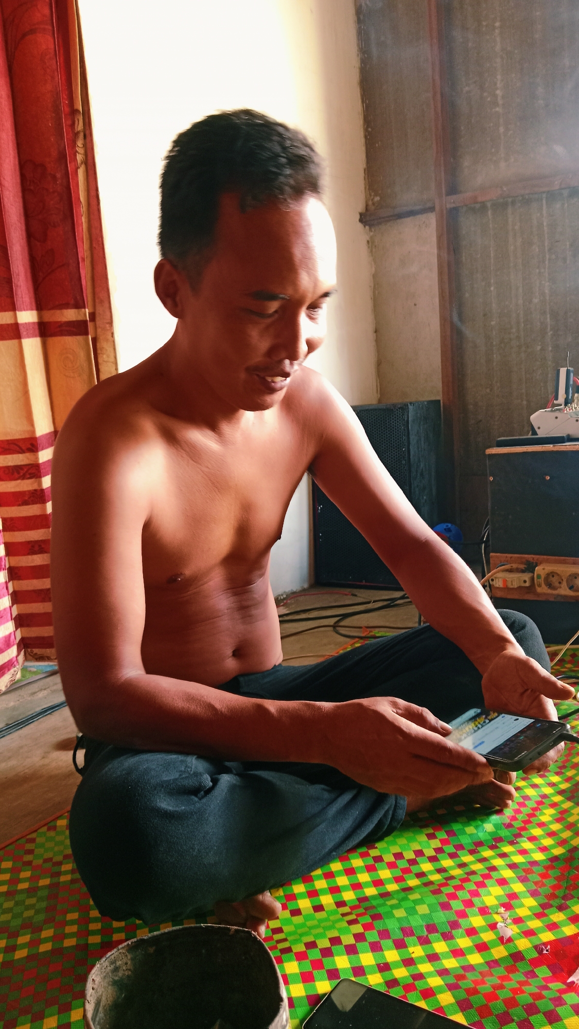 A shirtless East Asian man sits on a checkered mat in a dimly lit room. He is looking down at a smartphone in his hands. The man has short black hair and is wearing dark pants. In the background, there are large speakers, electrical outlets, and red and yellow curtains. A dark bowl sits to his left on the mat, and another smartphone is visible on the mat in front of him. The scene appears to be in a residential area of Karang Bunga, Indonesia.
