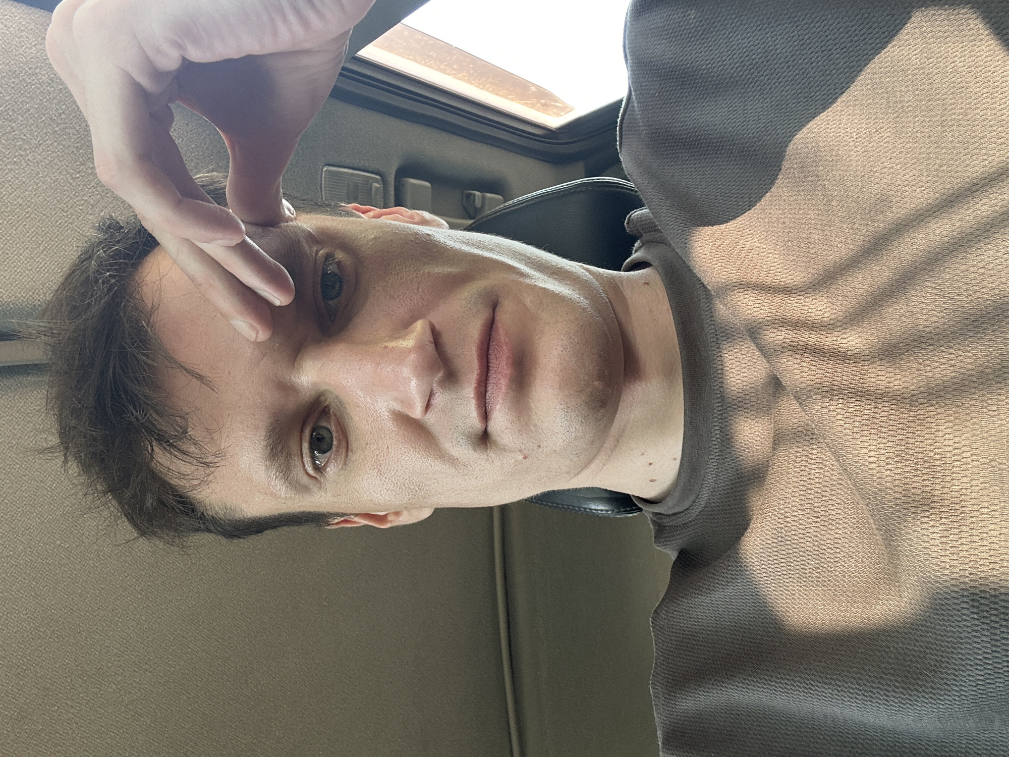 A man is sitting in the driver's seat of a car, looking into the camera. He has light skin, brown hair, and green eyes. He is wearing a gray t-shirt. His right hand is resting on his forehead. The interior of the car is visible in the background, with a headrest and part of the ceiling. Sunlight is streaming in from the right side of the car, casting shadows on the man's shirt and face. There is no indication of the city of Makiv, Ukraine, in the image.