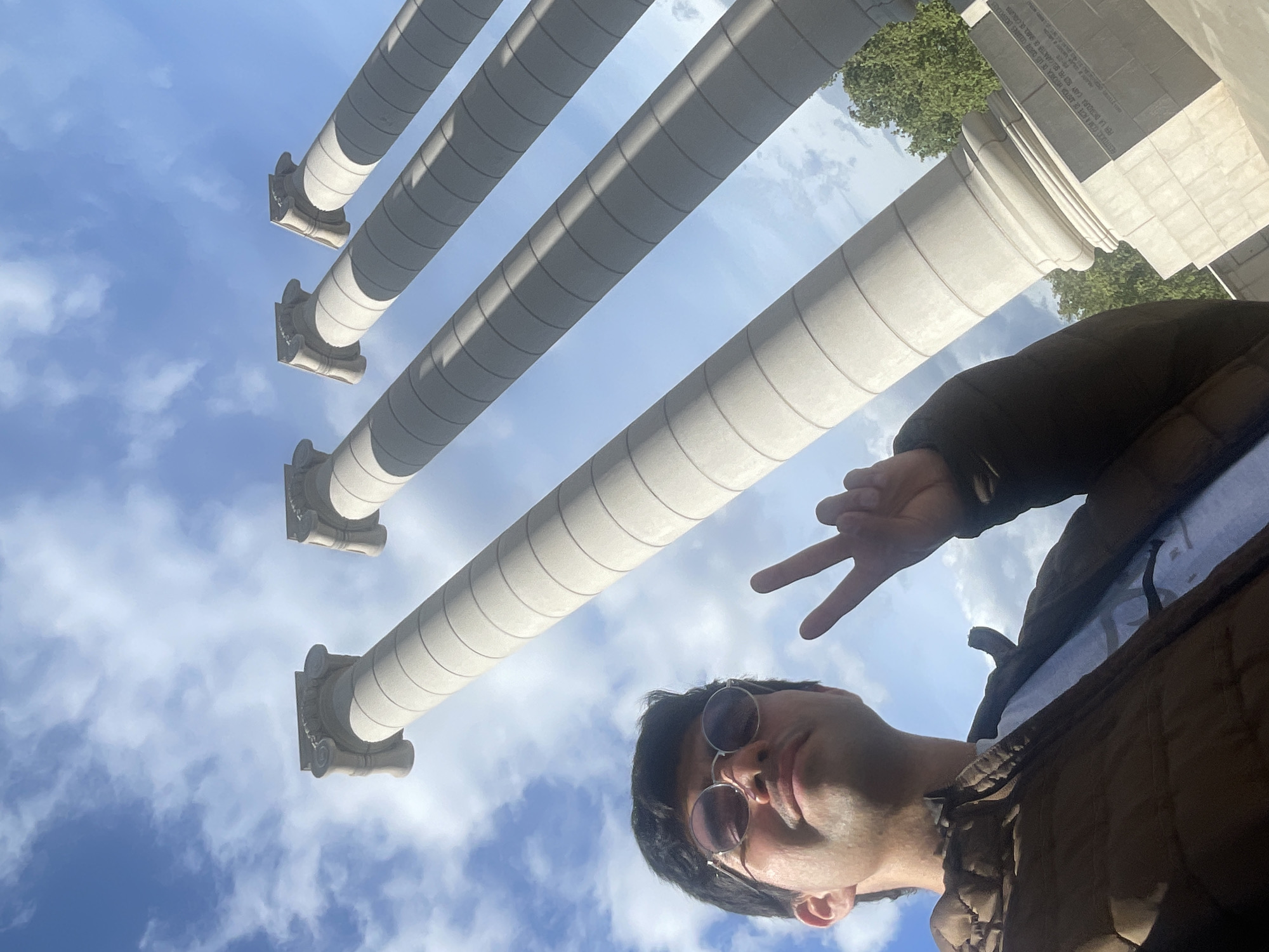 The image is a low-angle, upward-facing shot of a person standing in front of the Four Columns in Barcelona, Spain. The columns are large, gray, cylindrical structures with decorative tops and visible seams. The sky behind the columns is blue with scattered white clouds. The person is wearing sunglasses and a brown jacket and is making a peace sign with their hand. The scene appears to be taken during daylight hours, and a glimpse of greenery can be seen next to one of the columns. A stone base is visible beneath the columns, with lettering.