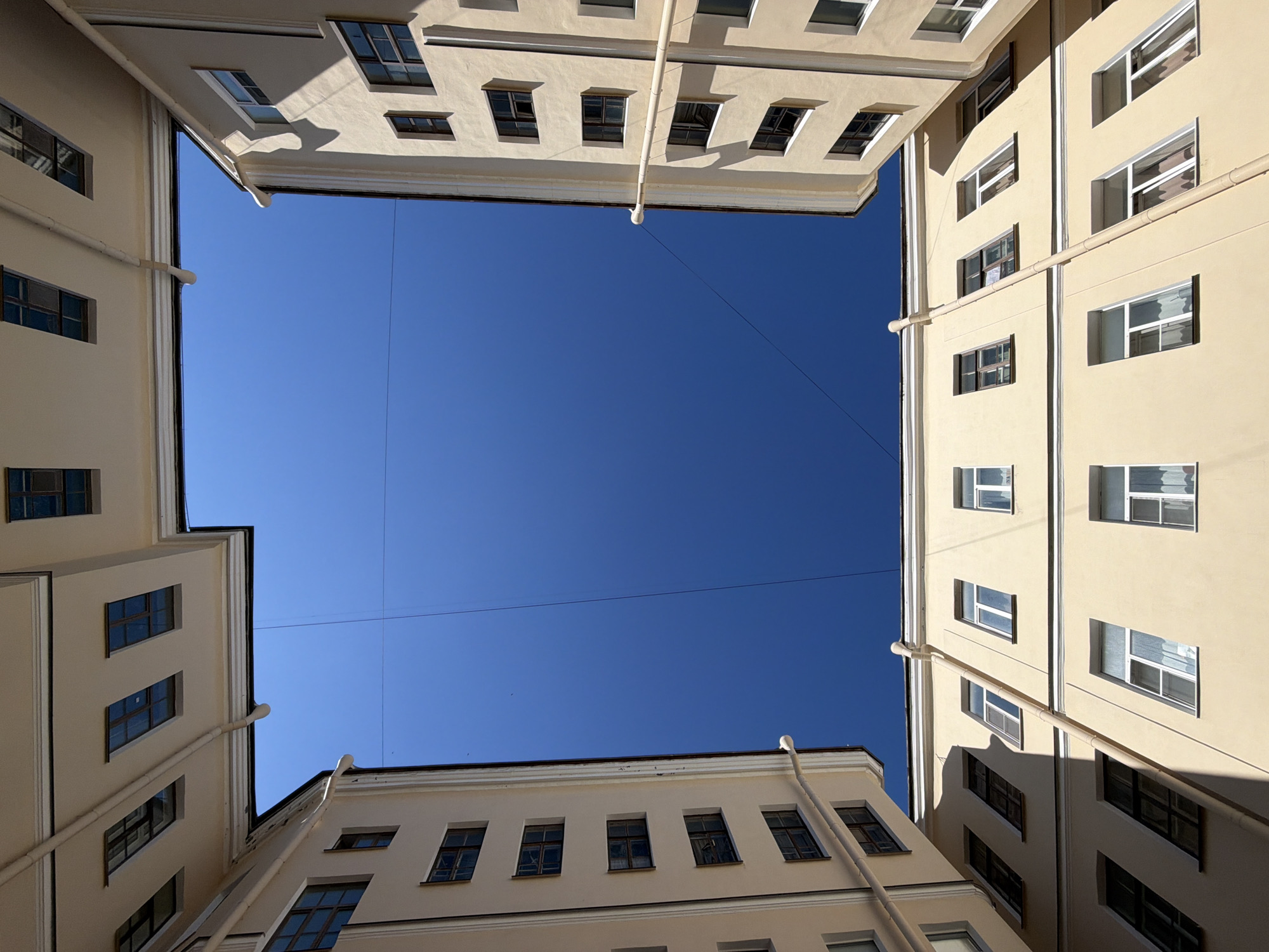 The image presents a worm's-eye view looking straight up from within a courtyard, likely in Saint Petersburg, Russia, framed by the surrounding buildings. Four sides of multi-story buildings, painted in a light yellow or beige color with prominent white architectural trim around windows and corners, converge towards the sky.
The central subject is a vast, clear, and bright blue sky, occupying the majority of the frame. Numerous rectangular windows are visible on the building facades; some are open, some closed, and many reflect the intense blue of the sky or distant light. White drainpipes run down the exterior walls of the buildings, adding vertical and diagonal lines to the composition.
The activity depicted is static; no people or specific events are taking place. The scene is characterized by its unique architectural perspective. Notable details include the bright, direct sunlight, which casts strong, crisp shadows on the building surfaces, particularly beneath cornices and window frames, indicating a clear, sunny daytime. Faint, thin lines or wires can be seen crisscrossing the blue sky. The architecture, with its warm-toned walls and distinct window styles, is characteristic of urban environments like Saint Petersburg. There is no visible text in the image.