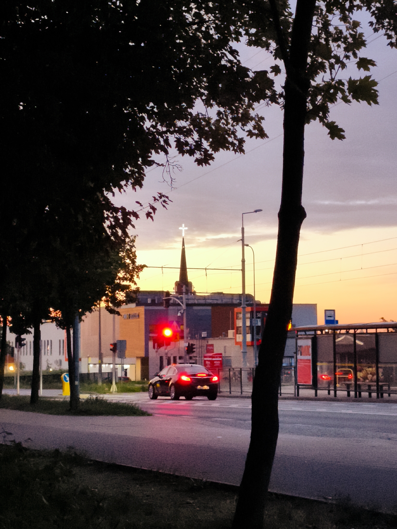 The image captures a street scene in Gdańsk, Poland, during twilight. A dark-colored car is stopped at a red traffic light, its brake lights illuminated. Across the street, a bus stop shelter is visible with a blue sign indicating a public transport stop. In the background, a church steeple topped with a cross stands out against the colorful sky, transitioning from orange at the horizon to purple and grey overhead. Several modern buildings with colorful facades line the street, and power lines are strung across the sky. Trees with dark foliage frame the foreground, with one tree trunk prominently positioned on the right side of the image. The overall atmosphere suggests an urban environment at the end of the day.
