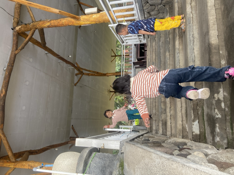 Two children are walking on outdoor steps. One, wearing a striped shirt and jeans, is mid-stride. Another, in a patterned shirt and yellow pants, is further up the steps. The steps lead past a building with wooden supports and a ladder. A millstone sits near the bottom of the frame. A stone wall borders the steps.