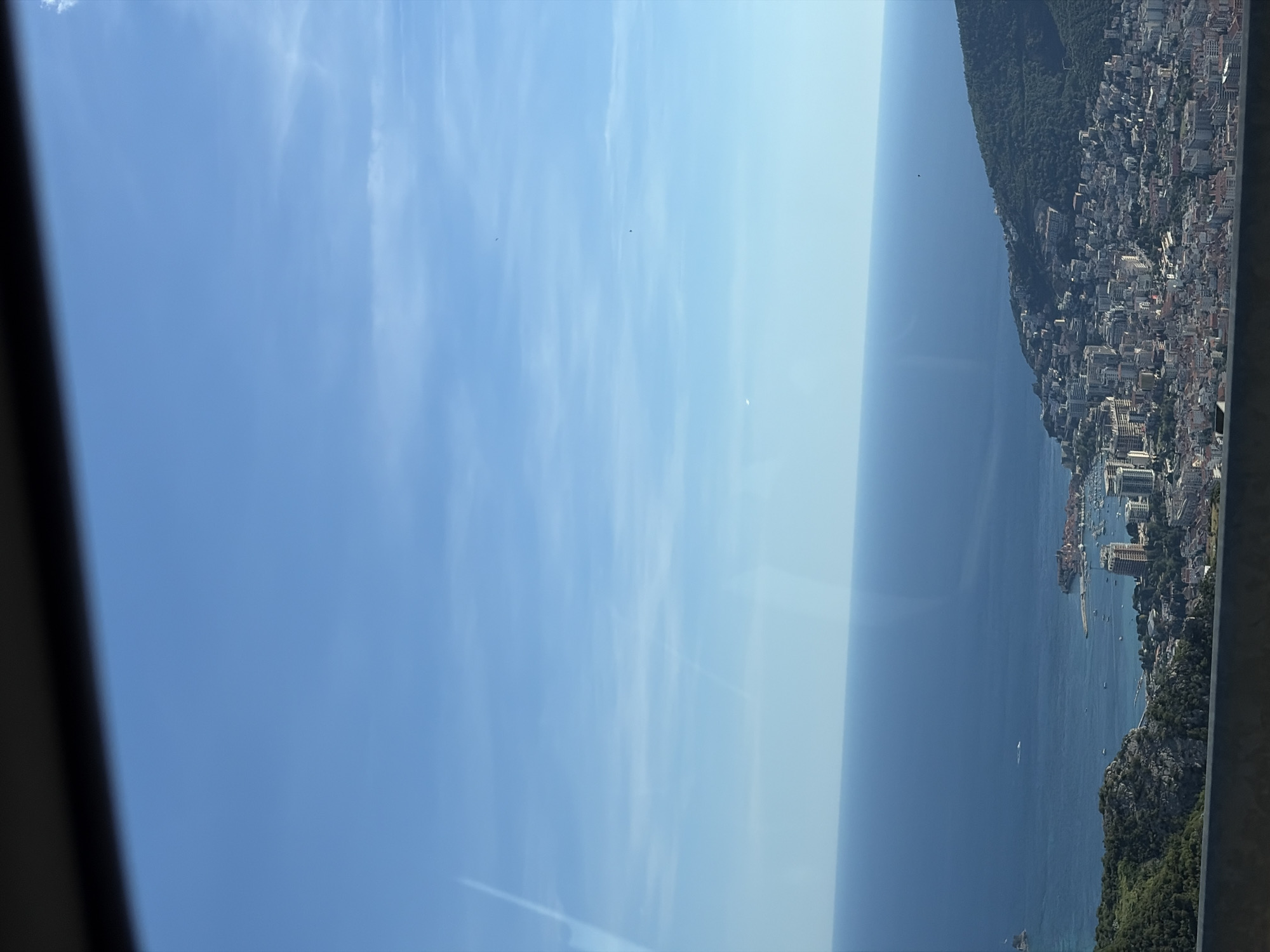 This image captures an elevated, panoramic view of the city of Budva, located in Montenegro. The scene is dominated by the vast expanse of the Adriatic Sea, stretching out to meet a clear blue sky dotted with faint wisps of clouds. In the foreground and mid-ground, the city of Budva unfolds along the coastline, characterized by a dense arrangement of buildings with terracotta rooftops and modern high-rises. A harbor filled with numerous boats and yachts is visible, along with a prominent peninsula featuring historical architecture. The cityscape is nestled against a backdrop of lush, green, mountainous terrain. The perspective suggests the photo was taken from a high vantage point, possibly from the surrounding hills or a higher elevation within the city.