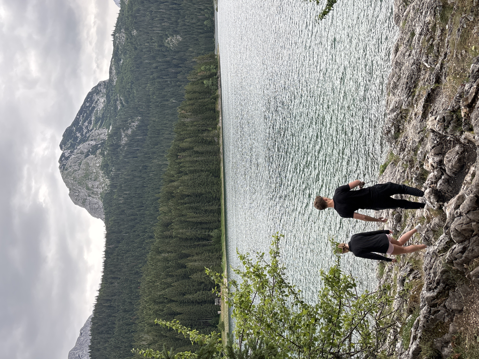 Two individuals, a male and a female, walk along a rocky shoreline next to a large, calm lake. The male, wearing a black t-shirt and black pants, walks slightly ahead of the female. The female, wearing a black long-sleeved top and pink shorts, walks behind and to the left of the male. The lake's surface is rippled, reflecting the overcast sky. Behind the lake, a dense forest of pine trees covers a mountain slope, leading up to a rugged, rocky mountain peak. In the far distance, more mountains are visible under a cloudy sky. On the left side of the image, a leafy green tree branch extends into the foreground. The environment suggests a natural, mountainous landscape, likely located in Pitomine, Montenegro, given the context.