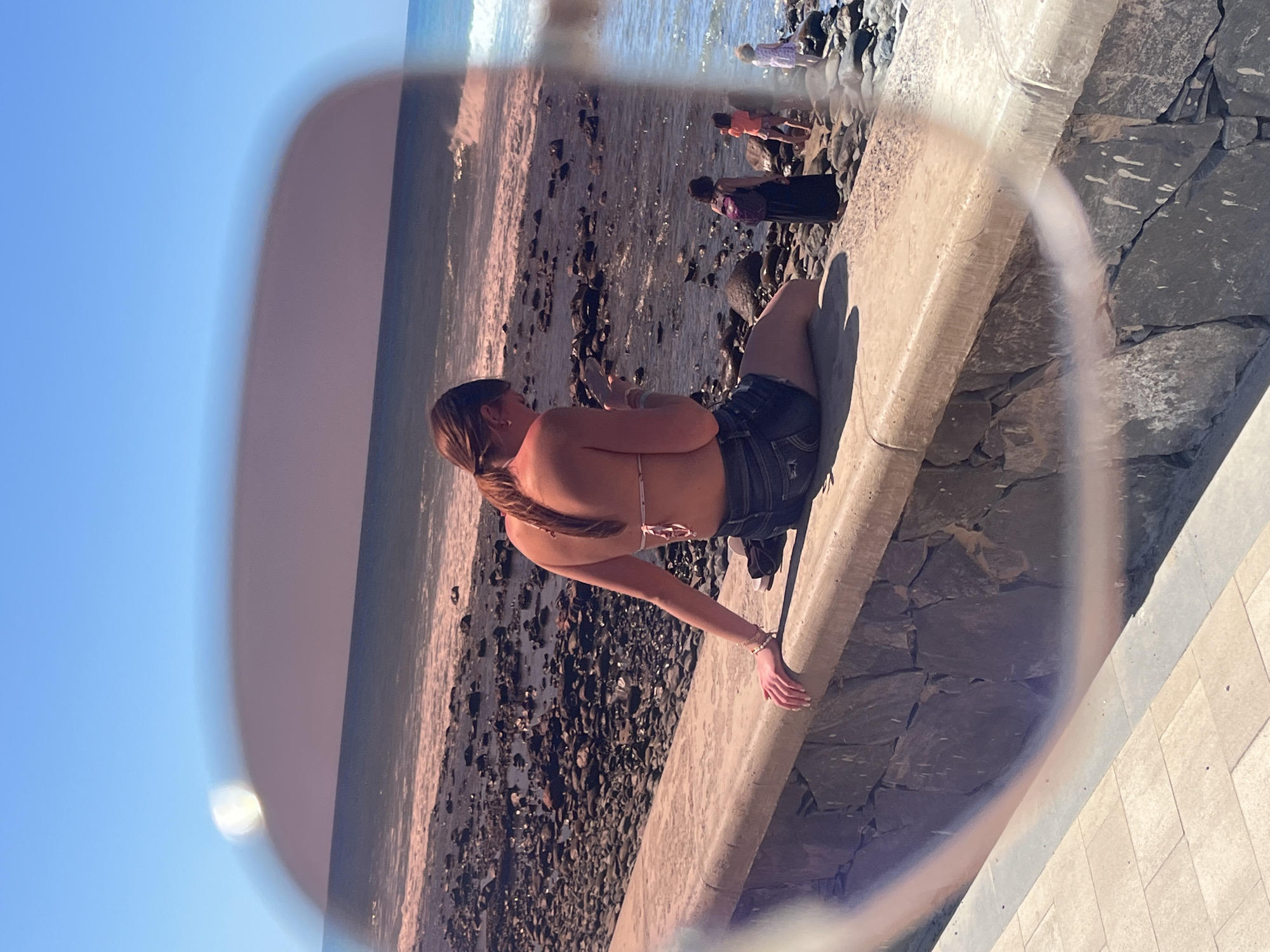 A medium shot, viewed through sunglasses, captures a beach scene in San Bartolomé de Tirajana, Spain. A light-skinned young woman wearing denim shorts and a bikini top sits on a concrete ledge, facing the beach. Her hair is pulled back in a ponytail. A beach with dark rocks and waves is visible. In the distance, several people are on the beach. The sky is blue and clear. The sunglasses frame partially obscures the scene, adding an artificial border to the image. The ledge is part of a wall constructed of stone and concrete.